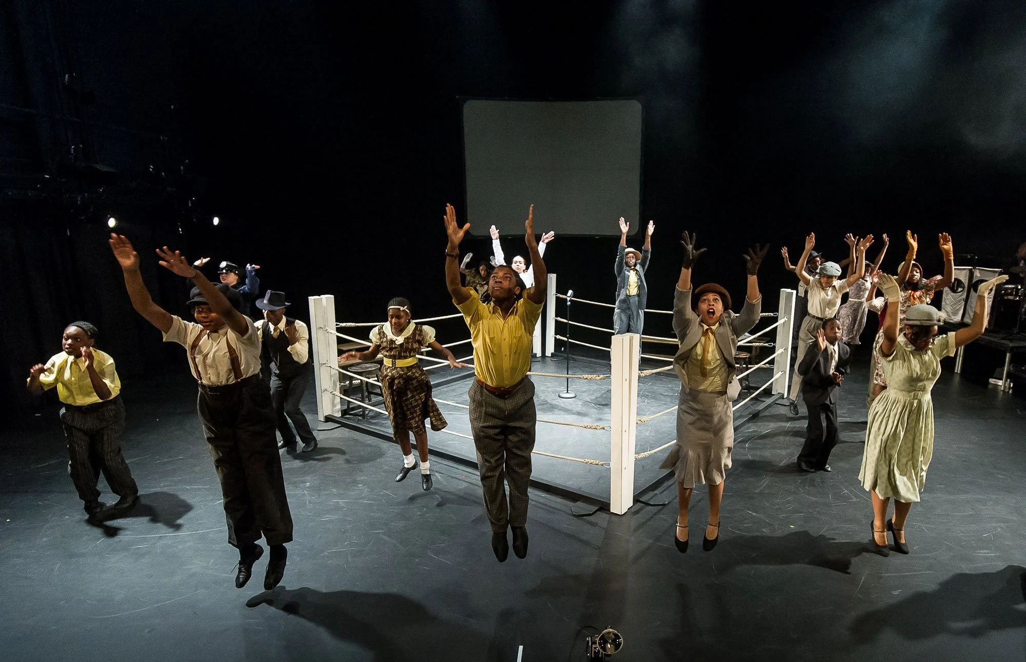 Actors performing a scene with a boxing ring, some actors are jumping and others are standing with raised hands.