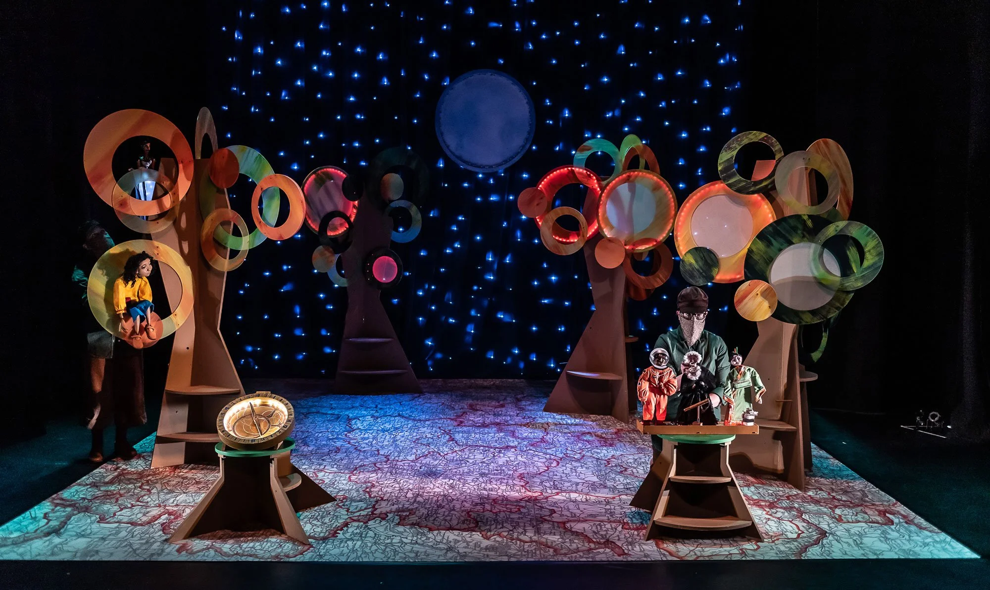 Stage with fantasy trees, glowing blue lights, a clock, and puppeteers operating puppets, set in a theatrical scene.