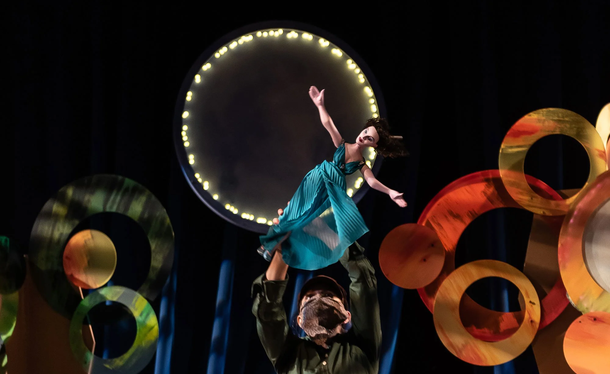 A puppet of Heddy Lamarr being lifted on a stage with colourful abstract circular decorations and bright background lighting.