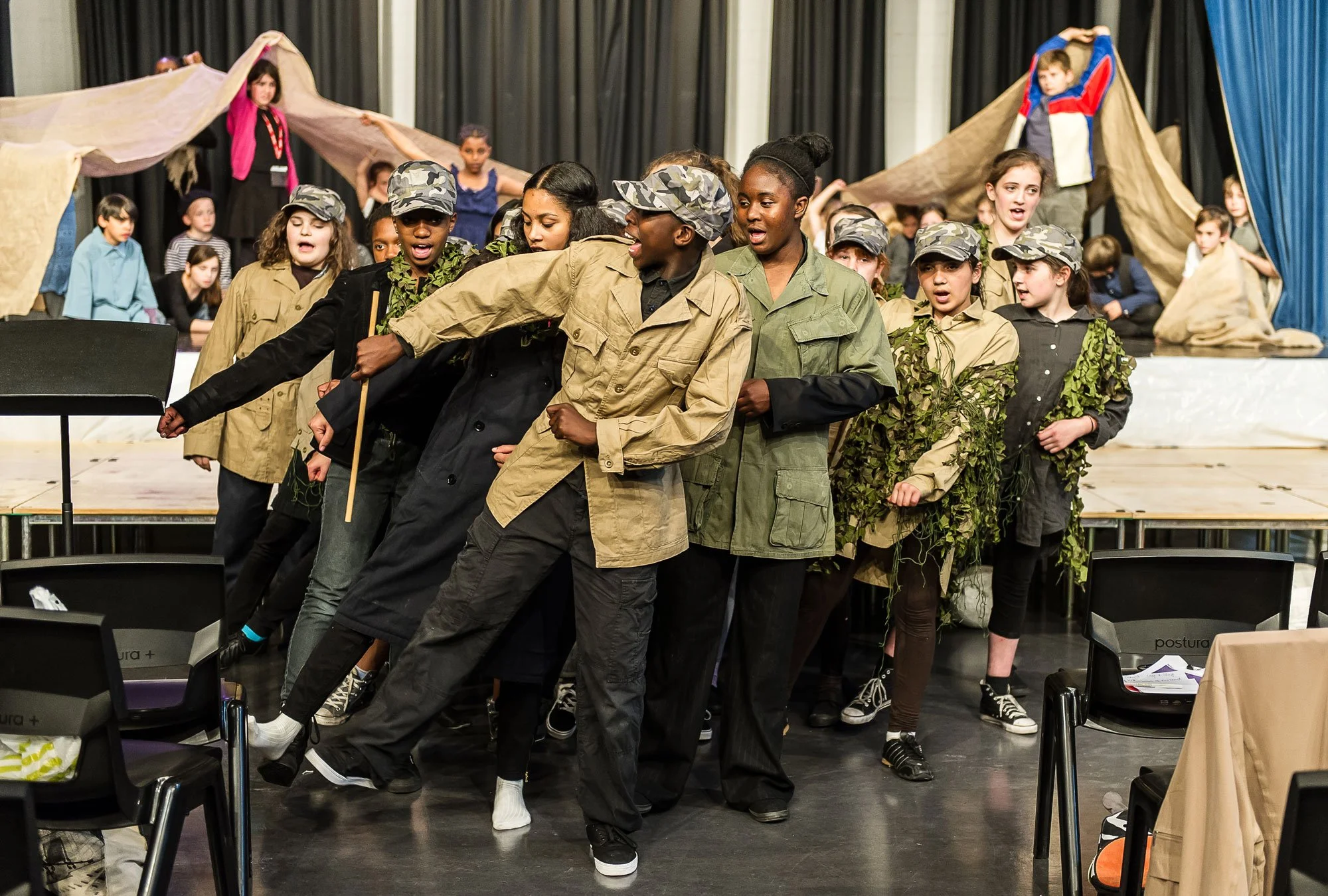 Children performing in a play, dressed in costumes resembling soldiers or explorers, with some wearing camouflage and others in costumes with leaves, on a stage with a backdrop depicting tents and a rocky landscape.