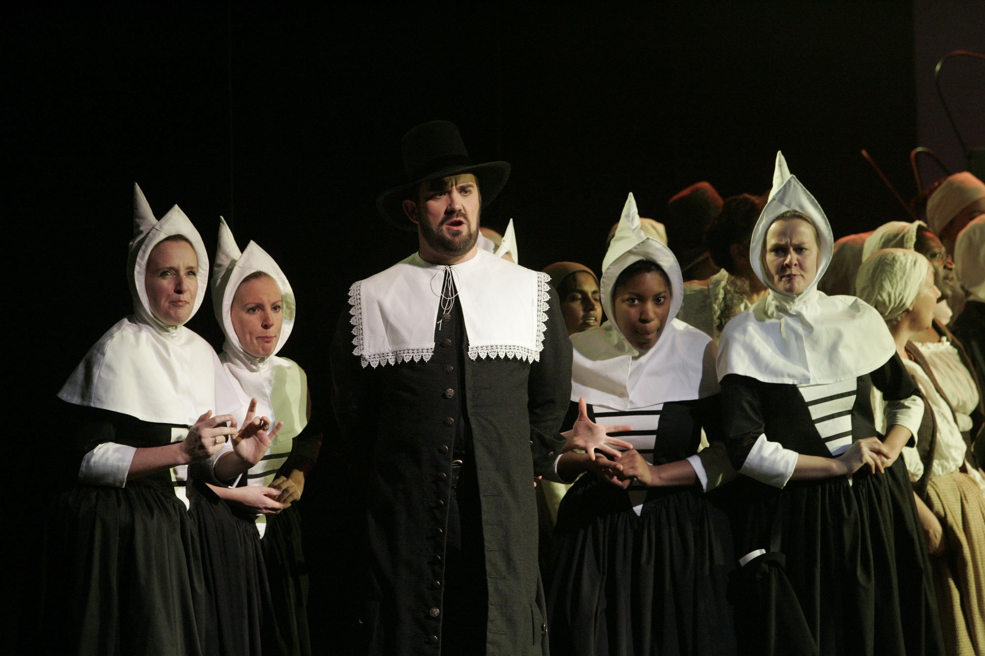 Theatrical performance with actors in historical costumes, including women in white bonnets and black dresses, and a man in a black coat and hat, on stage.