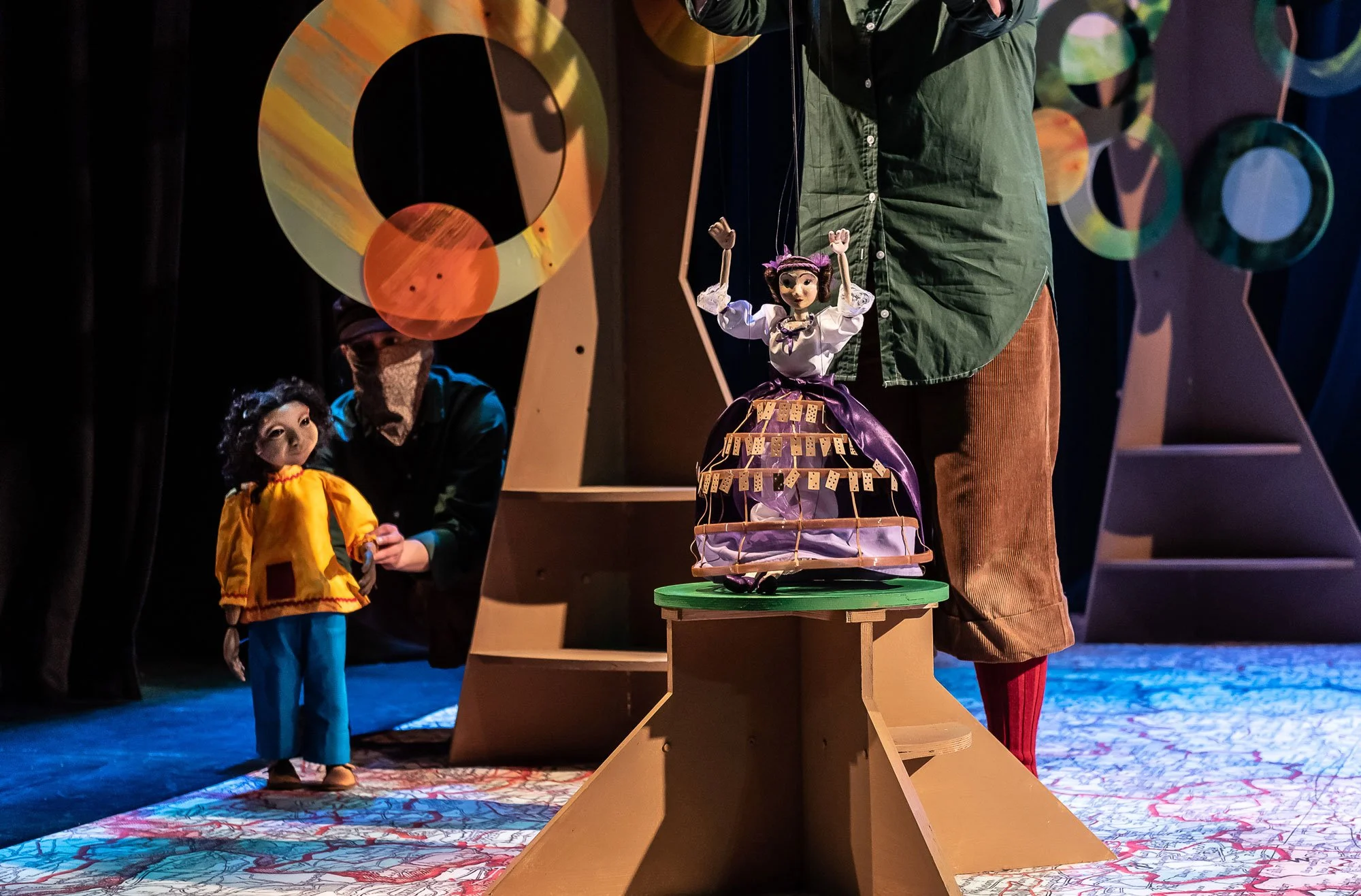 A puppet show scene featuring a puppet of Ada Lovelace standing on a platform, with two puppeteers.