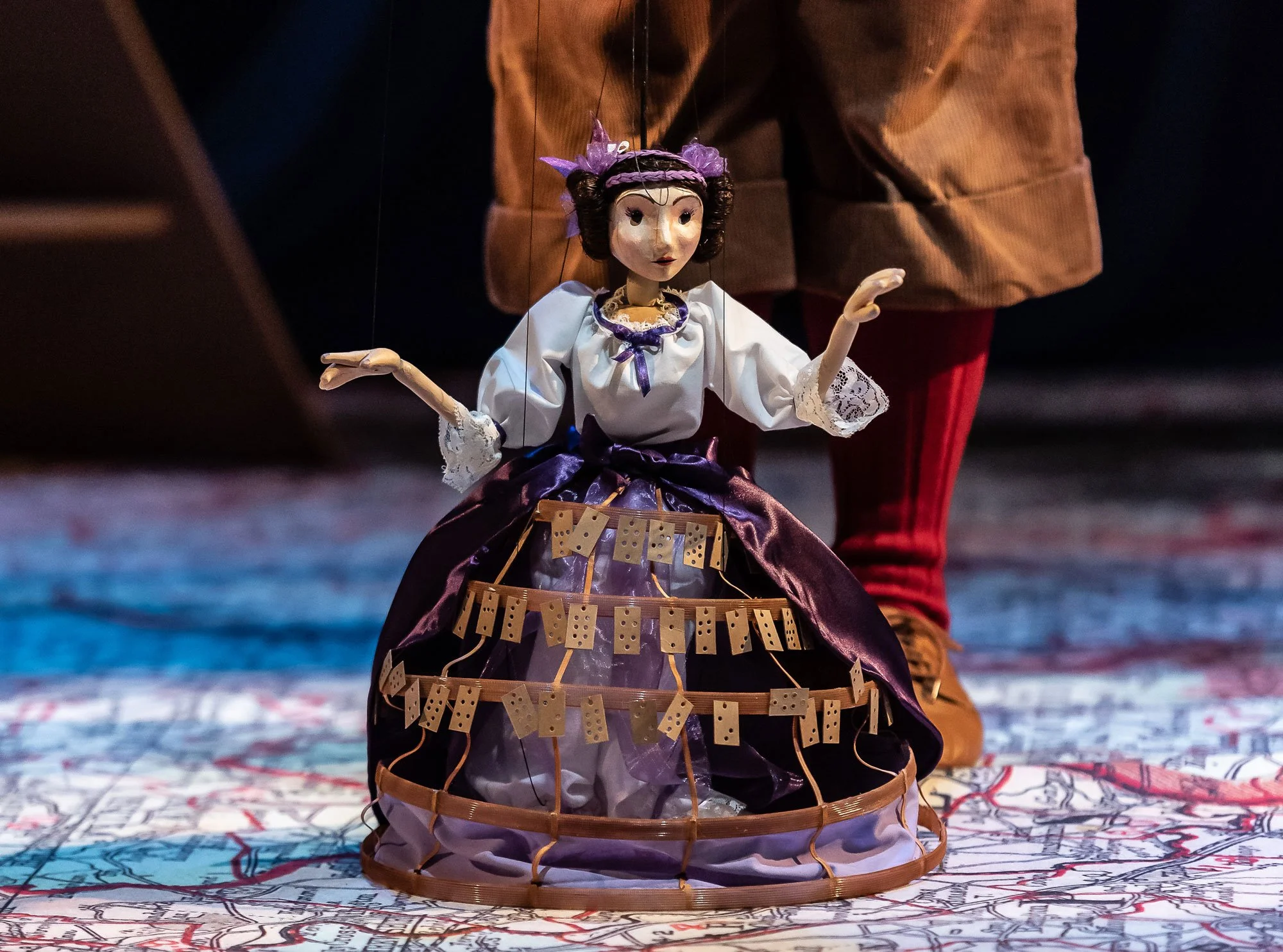 A marionette puppet of Ada Lovelace with a detailed dress and a cage-like skirt, is manipulated on a stage with a map in the background.