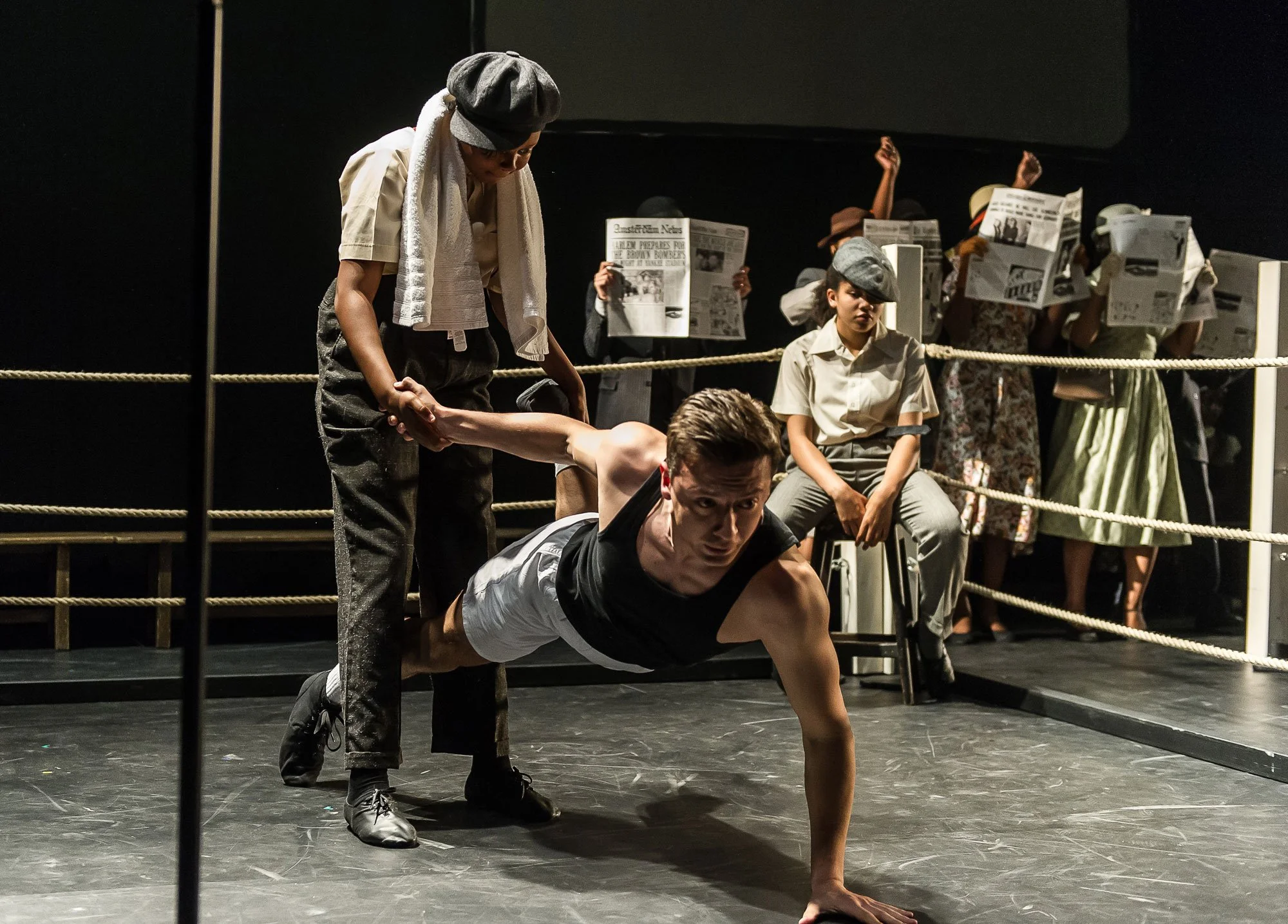 A theatrical scene with a man doing a push-up, holding a hand of a woman dressed in vintage clothing, with women in period costumes and newspapers behind a stage barrier.