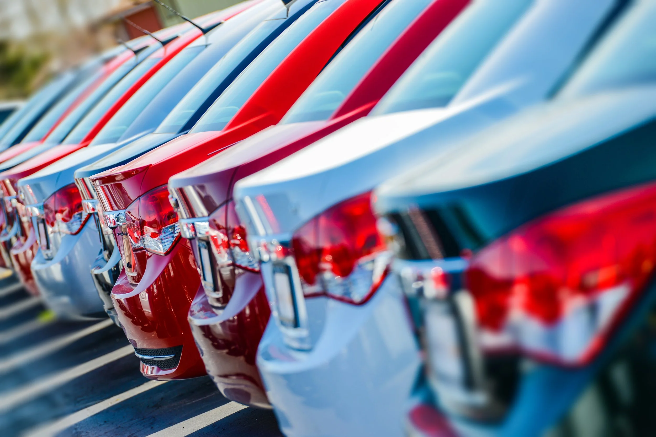 Lineup of parked cars in red, light blue, and green, viewed from the rear, in an outdoor parking lot.