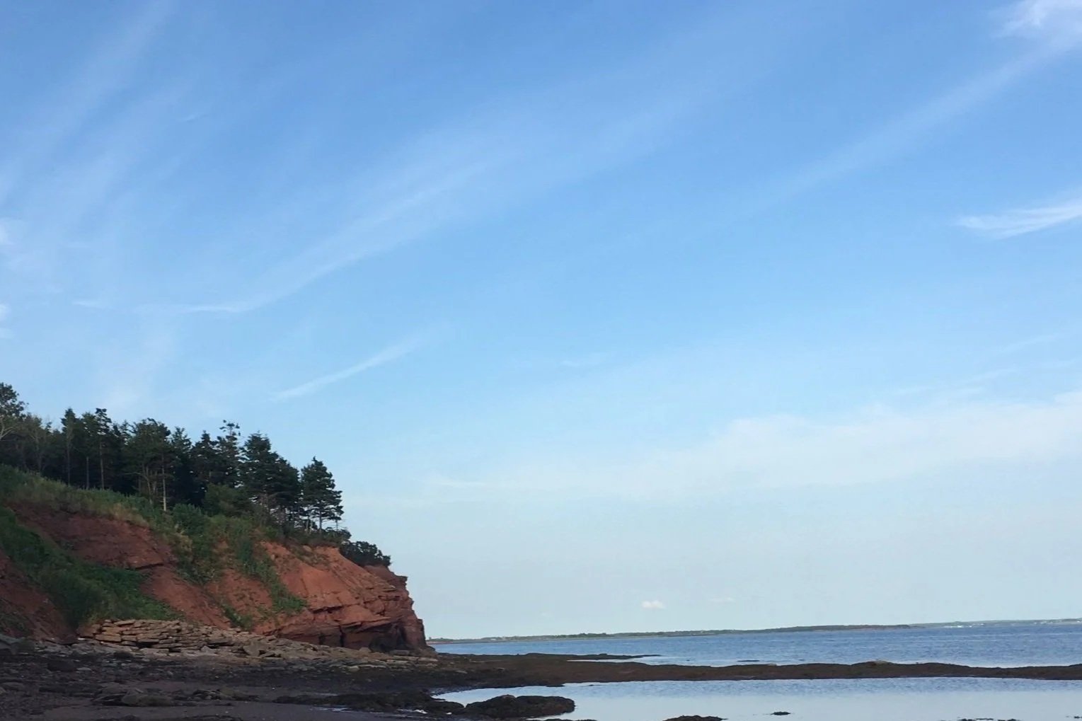 A coastal scene with a red cliff covered in trees on the left, a calm body of water, and a blue sky with wispy clouds.