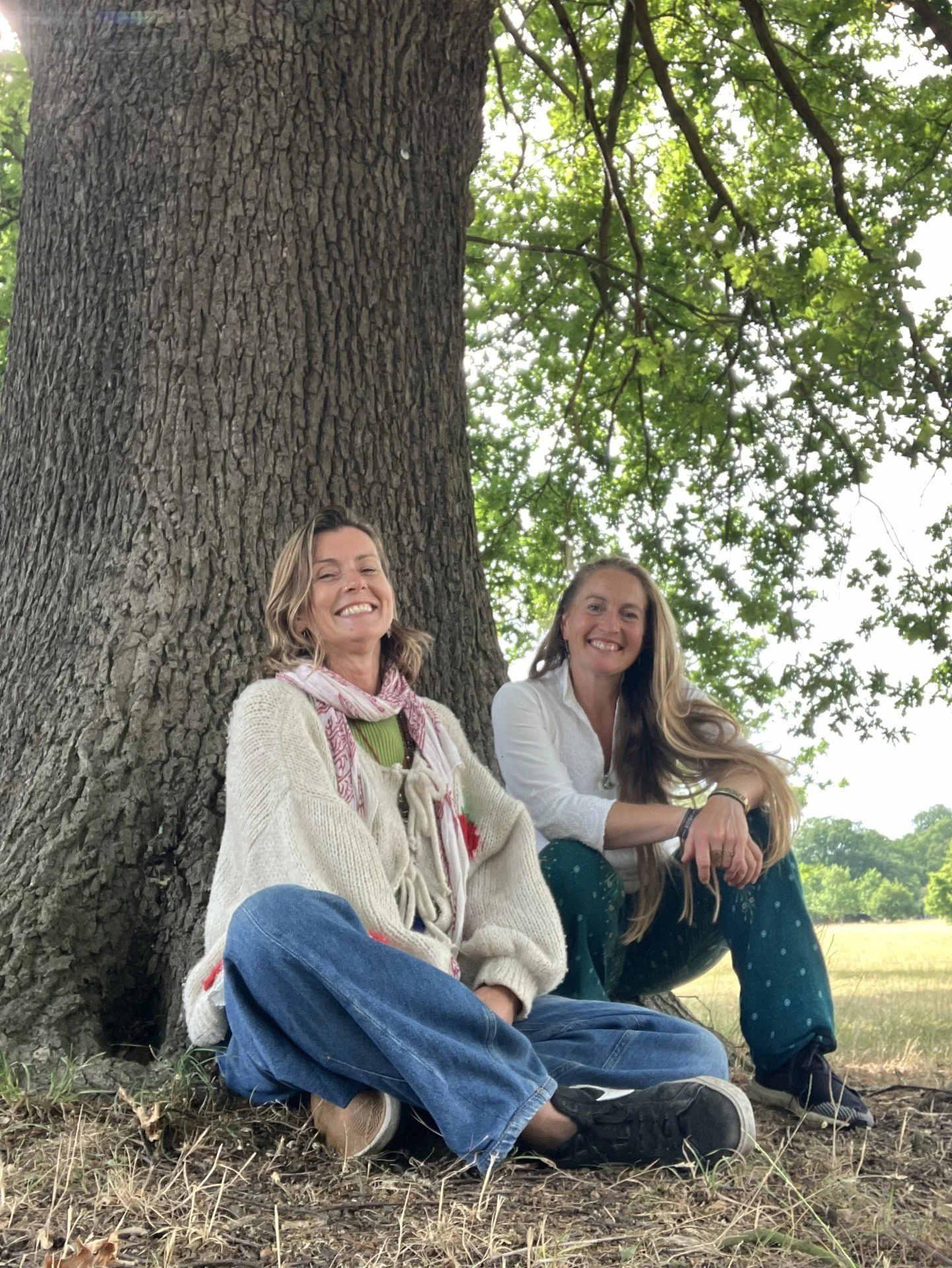 Two women sitting under a large tree, smiling, with green leaves overhead and an open field in the background.