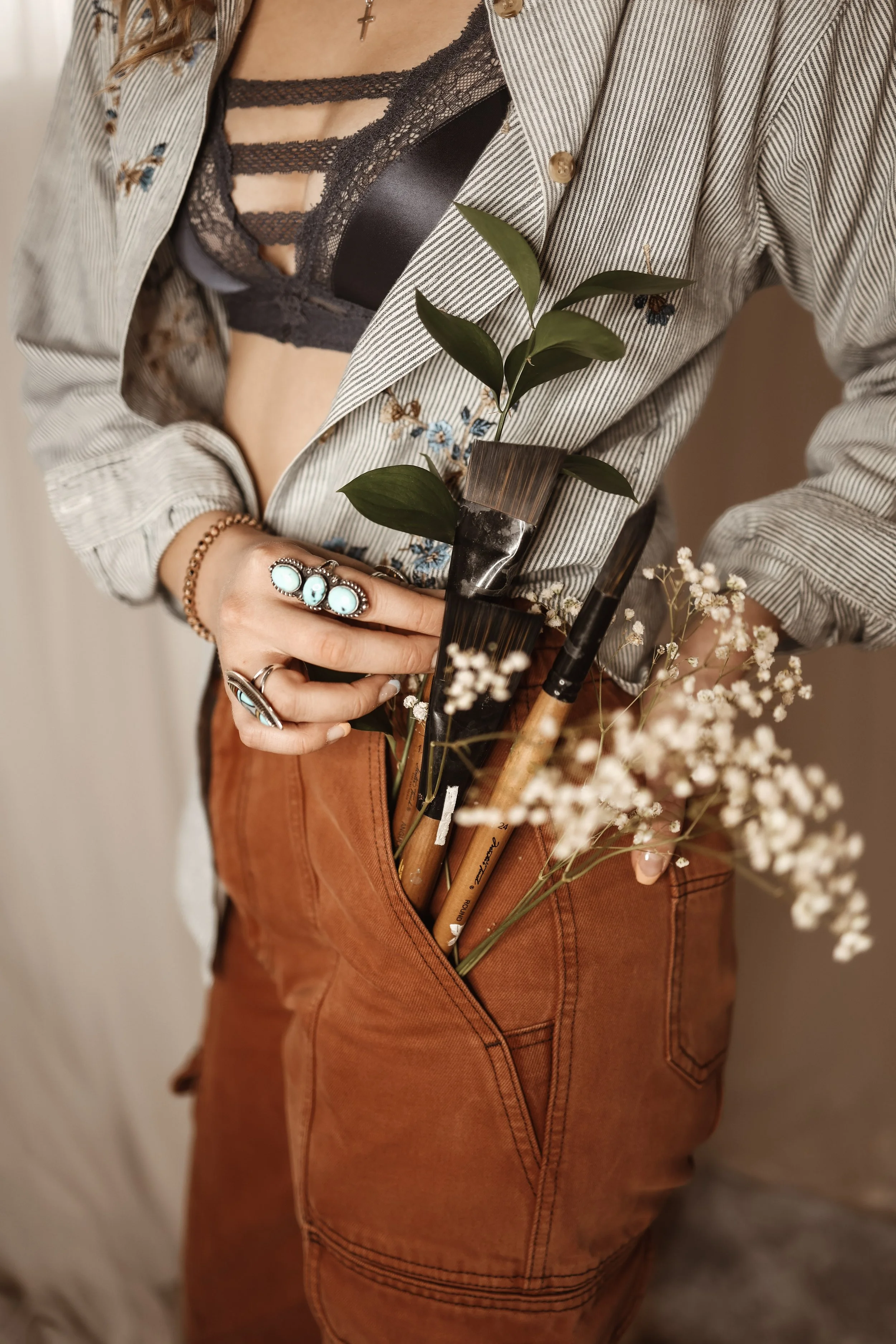 Person wearing a striped shirt with embroidery, black lace bralette, and rings on the fingers, holding a pot of green leaves and flowers while standing in front of a plain background.