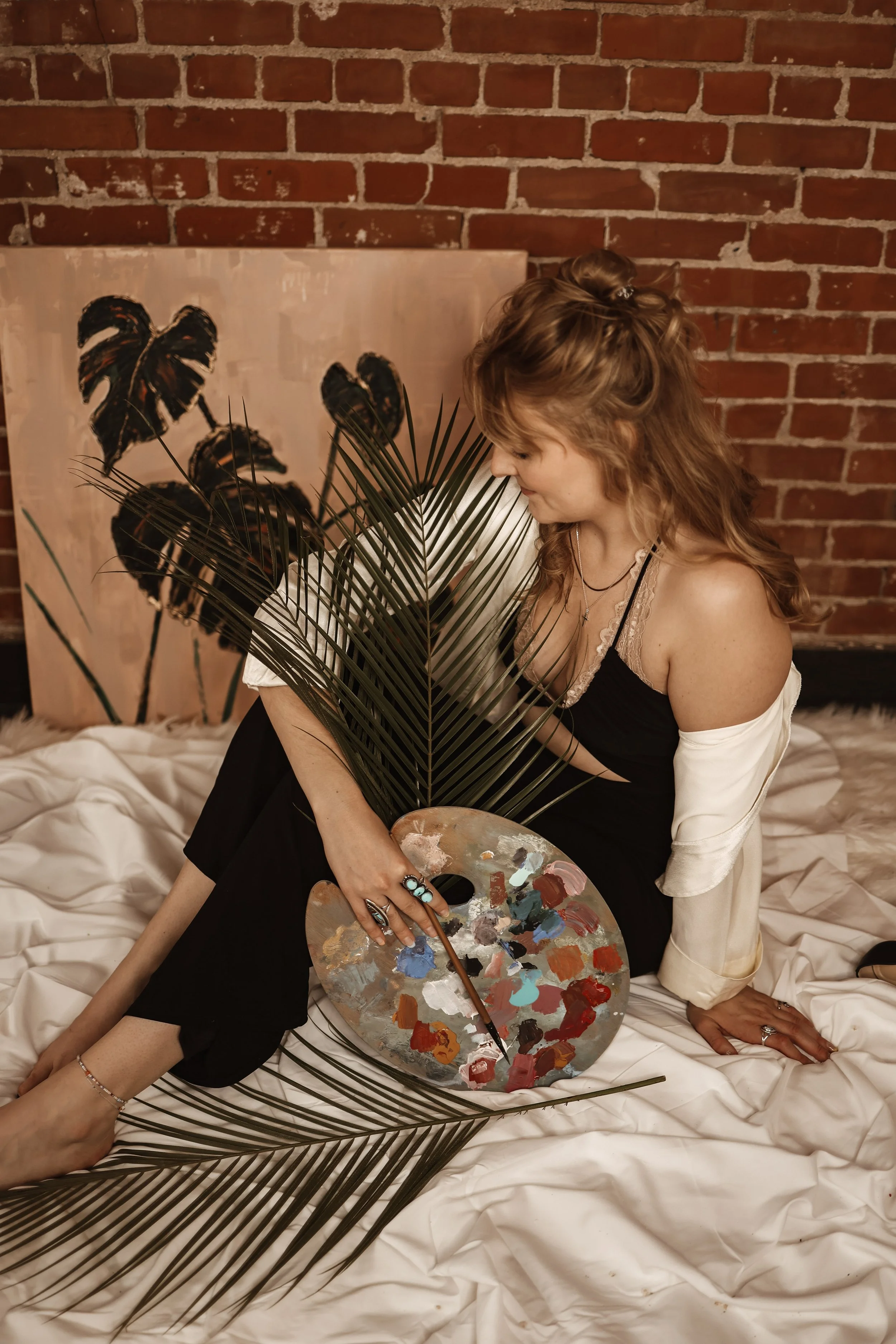 A woman with curly red hair, wearing a black dress with lace straps and a beige off-shoulder cardigan, is holding a painter's palette with colorful paints and a paintbrush. She is sitting on a bed covered with white sheets, with a large painting of dark tropical leaves leaning against a brick wall behind her. She is partially obscured by a large palm leaf in front of her.