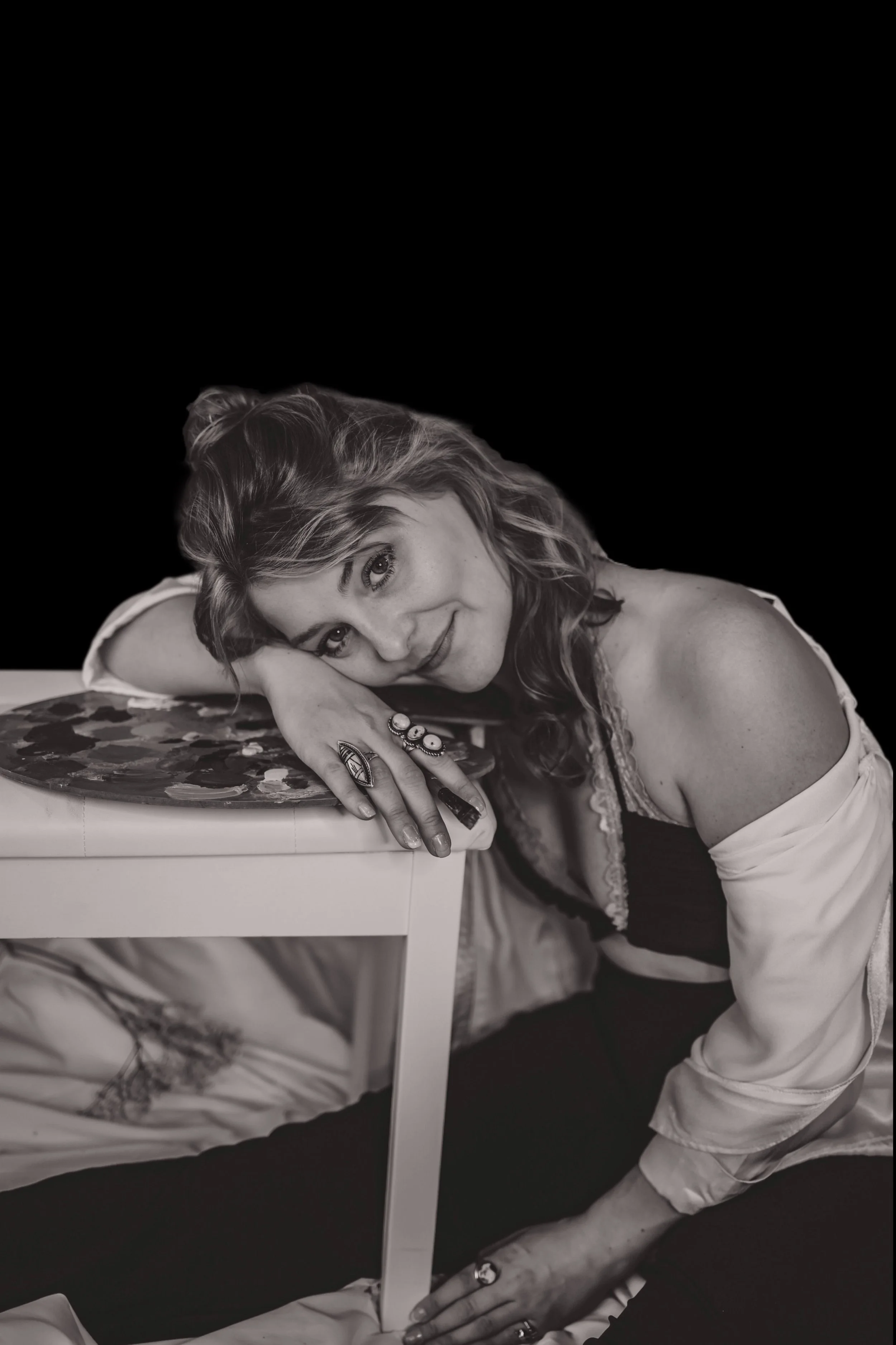 A woman with wavy hair, wearing rings, lying on her side with her head resting on her arm, smiling at the camera. She is leaning on a piece of furniture, wearing a white jacket over a dark top, with a dark background.