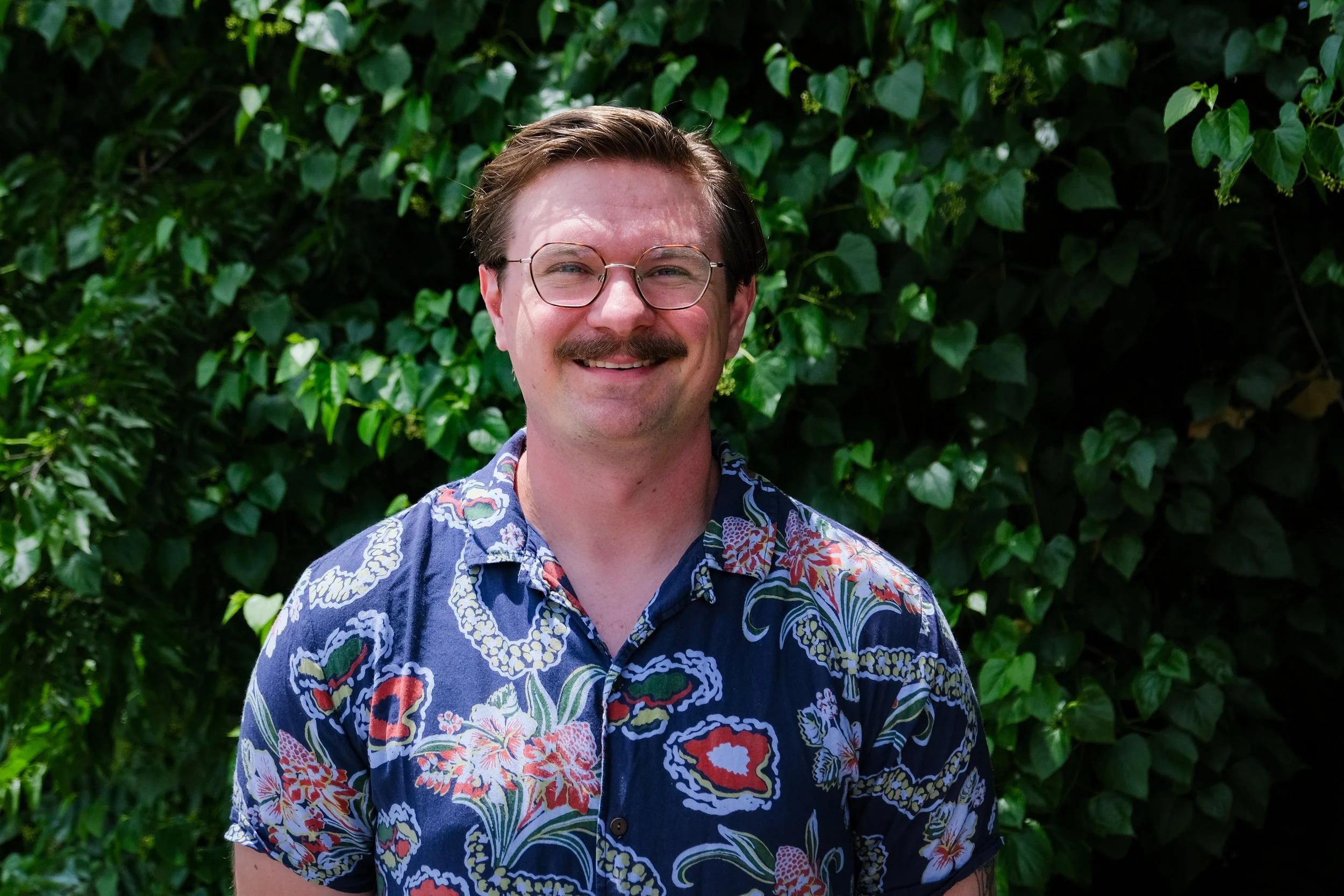 Gavin is a white man with glasses and a mustache. He is wearing a blue and floral printed button down.