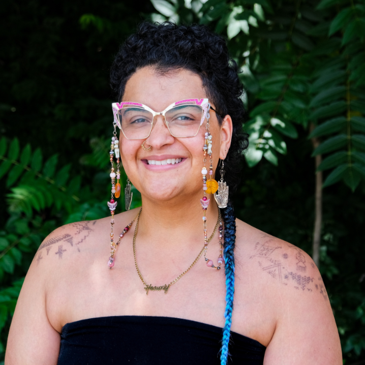 Cory is a Black woman with very colorful glasses, a long blue braid, and tattooes on her shoulders