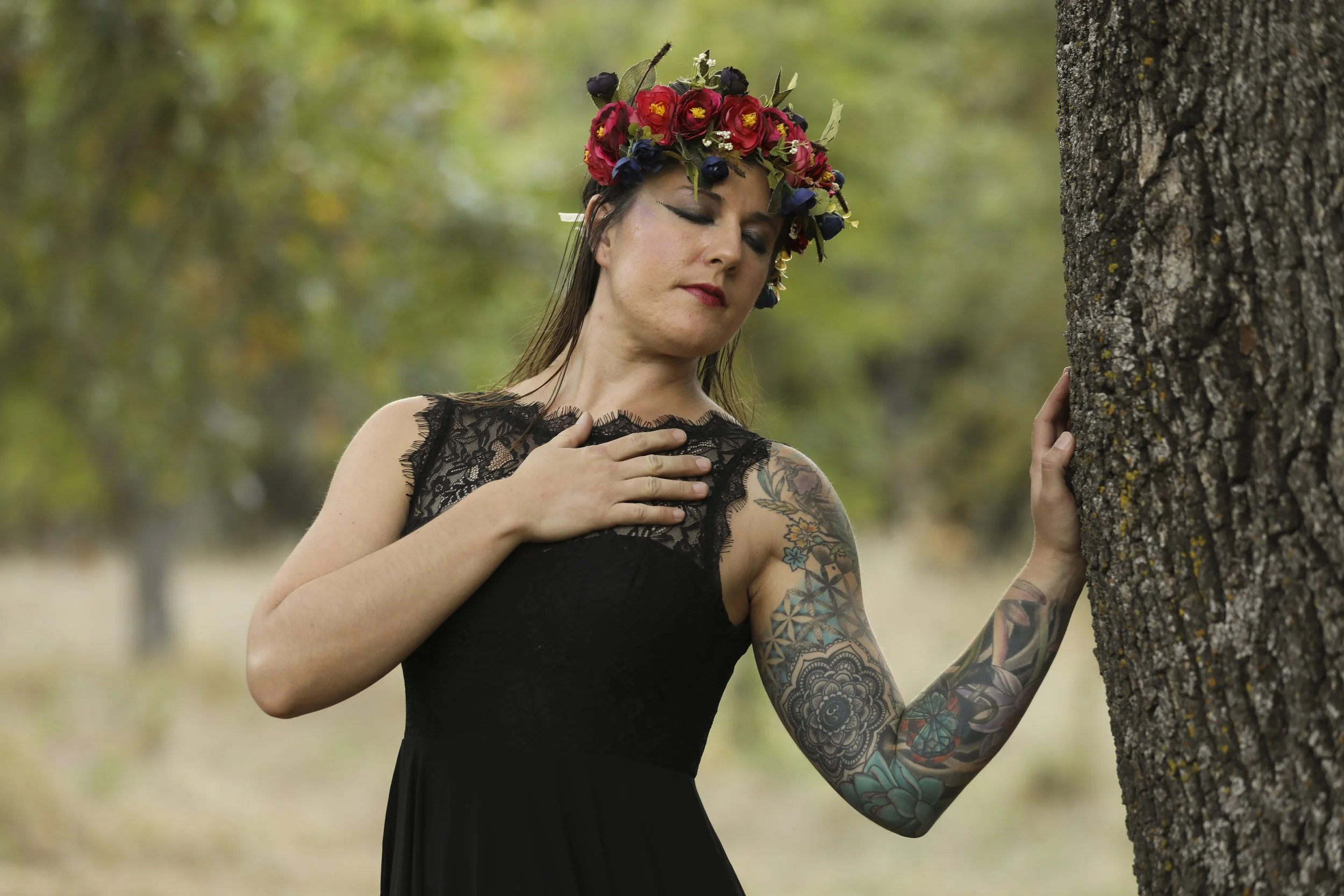 A woman wearing a black dress and a colorful flower crown, with tattoos on her arm, stands outdoors with her hand on a tree and her eyes closed.