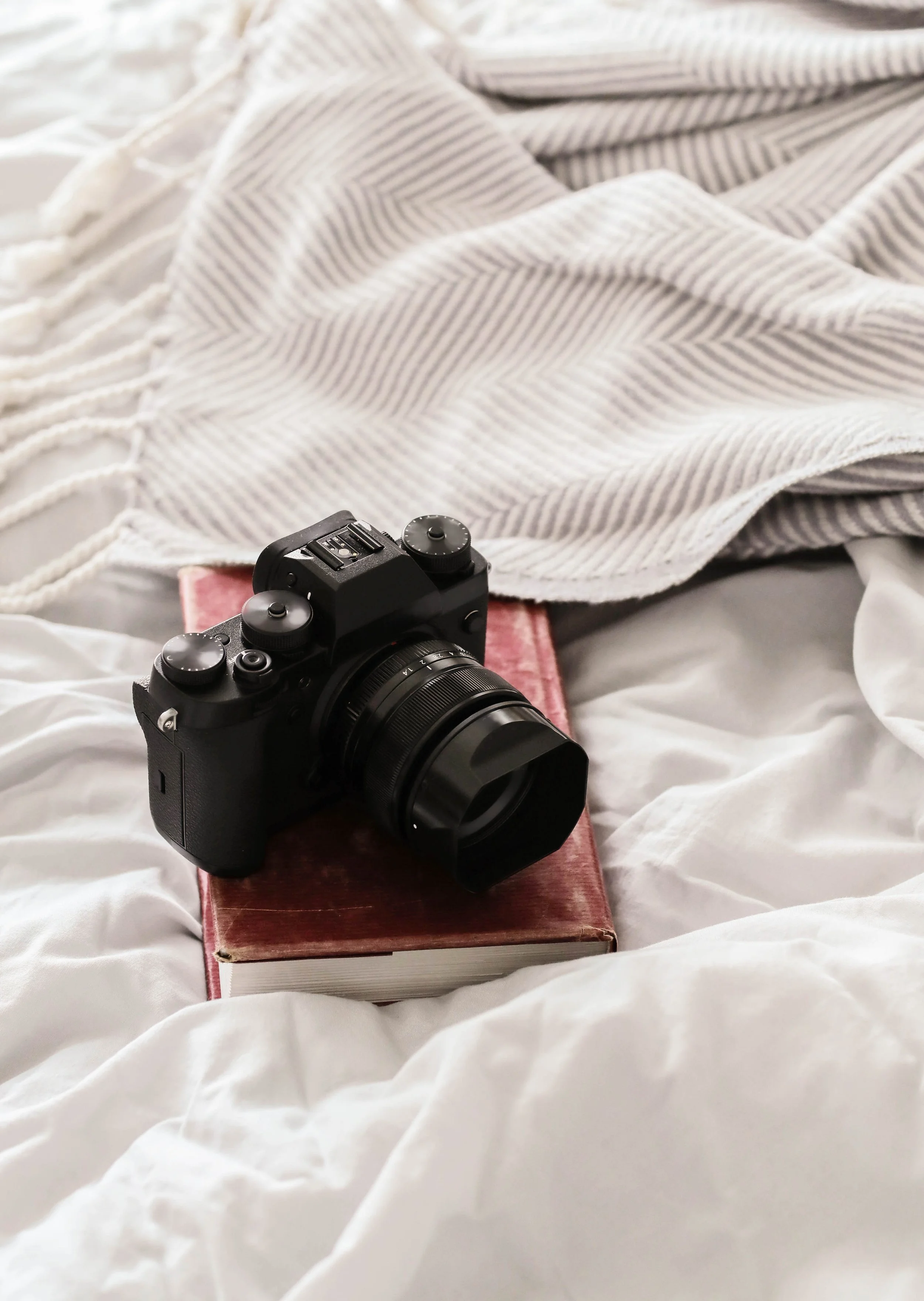 A black camera with a large lens resting on a closed red book, with a white and gray striped blanket in the background and white bedding underneath.