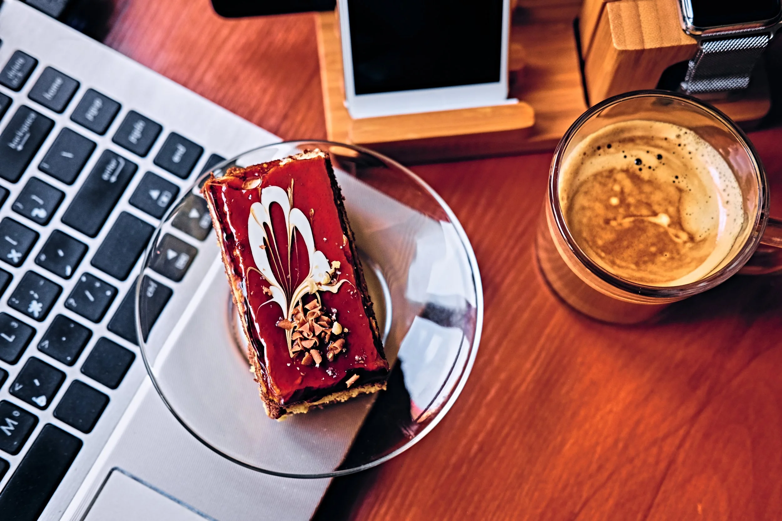 Laptop, a slice of red and gold patterned cake, a glass of coffee, and a smartphone on a wooden table.