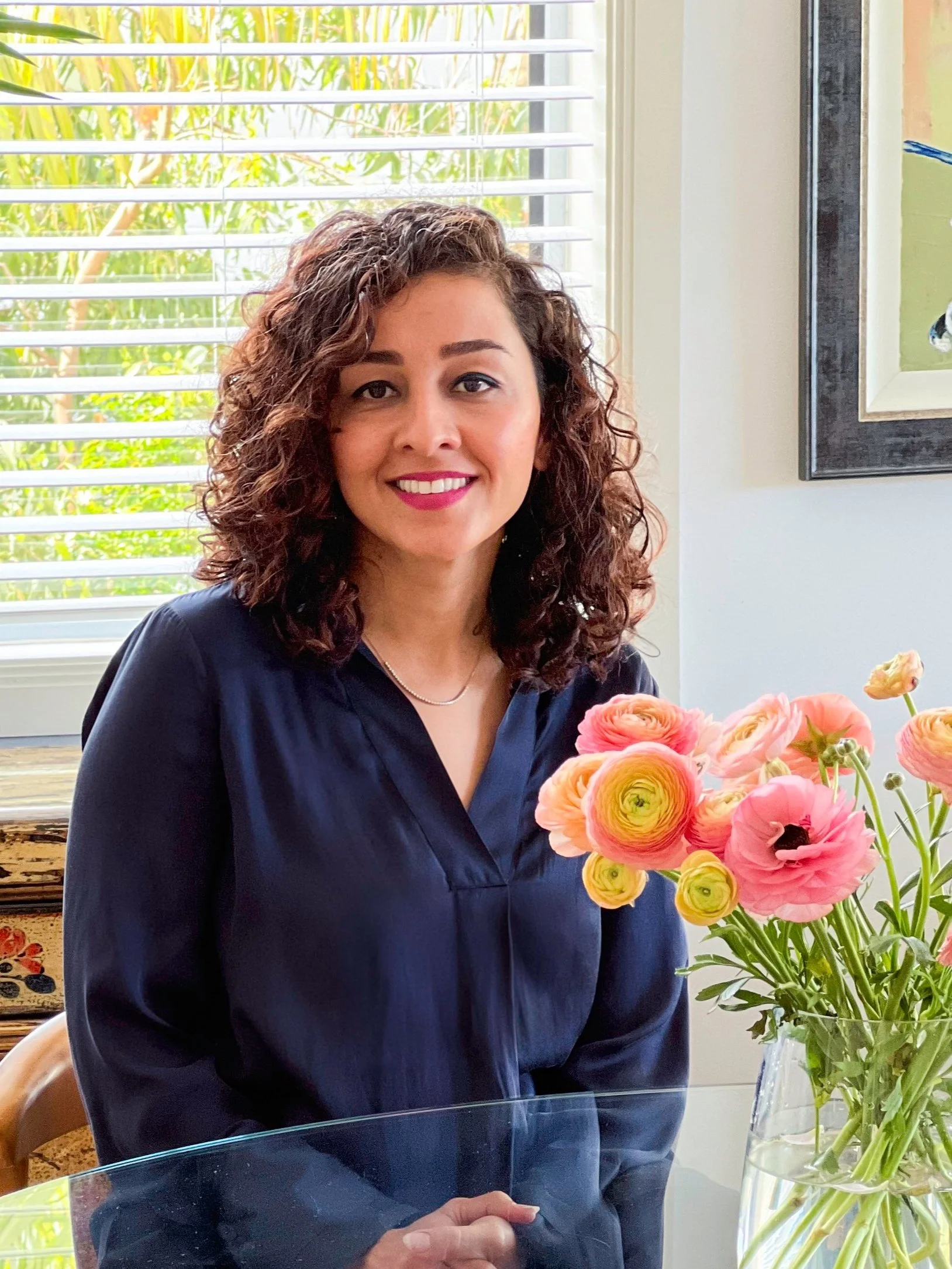 Ensieh Noorian A smiling woman with curly brown hair wearing a navy blouse sitting at a table with a vase of pink and yellow ranunculus flowers.