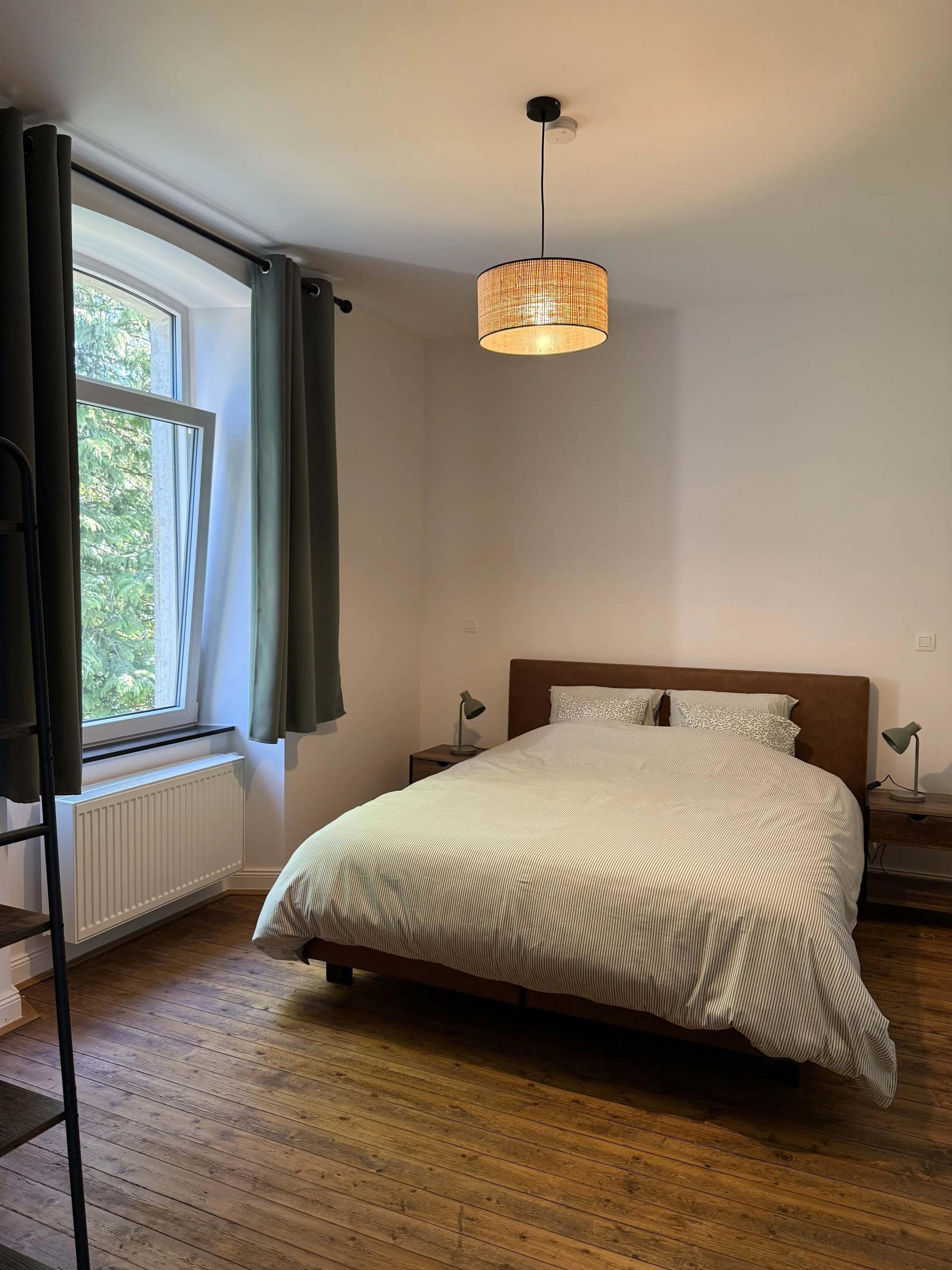 Chambre avec grand lit, deux lampes de chevet, fenêtres avec rideaux gris, parquet en bois, lampadaire suspendu en rotin au plafond.