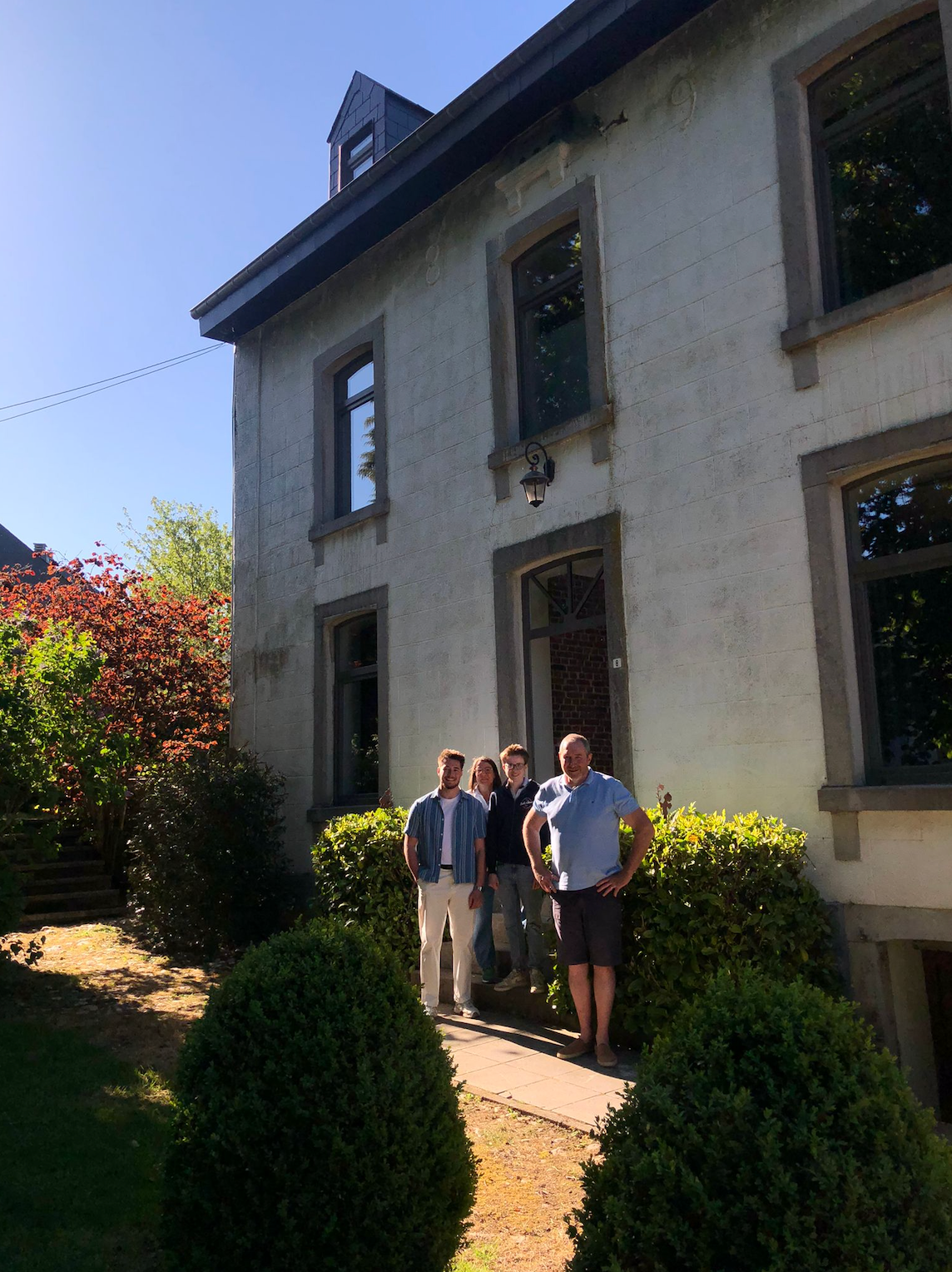 Quatre personnes debout devant une maison en pierre avec de grandes fenêtres et un jardin bien entretenu par un jour ensoleillé.