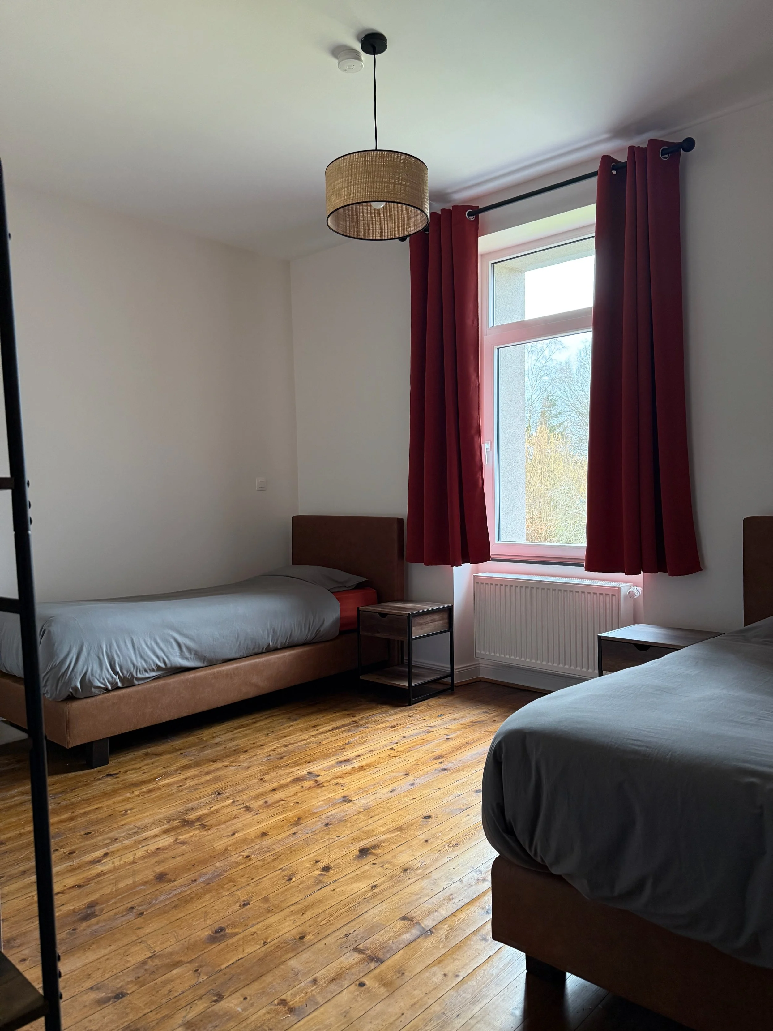 Deux lits simples dans une chambre avec parquet en bois clair, une fenêtre avec rideaux rouges, et un luminaire suspendu au plafond.