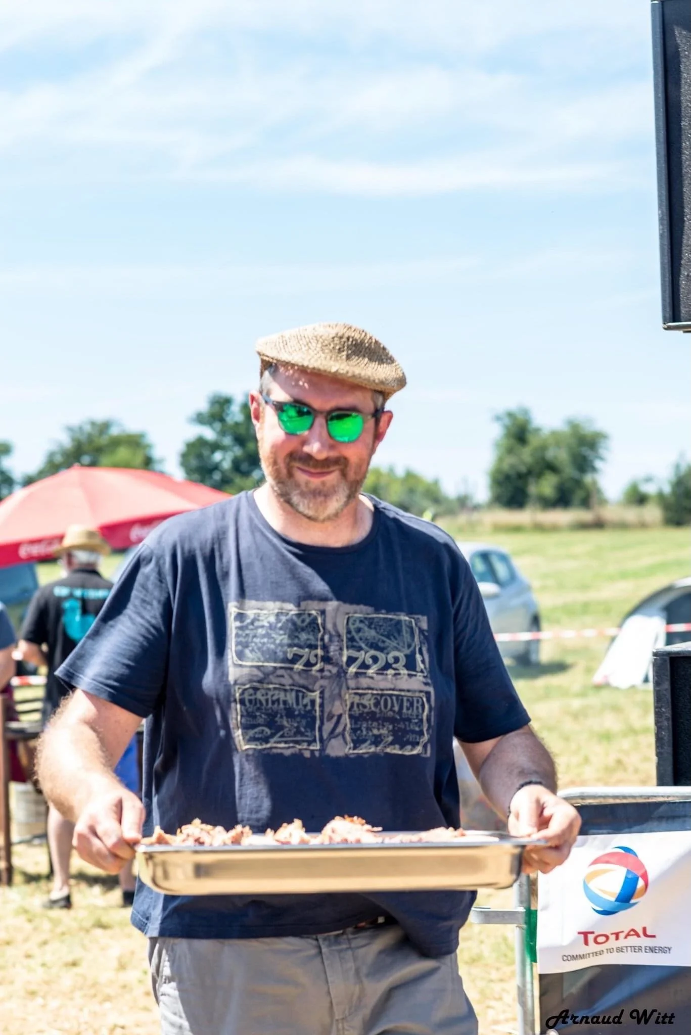 Un homme portant un chapeau en paille et des lunettes de soleil vertes, tenant une plaque de viande grillée lors d'un événement en plein air ensoleillé