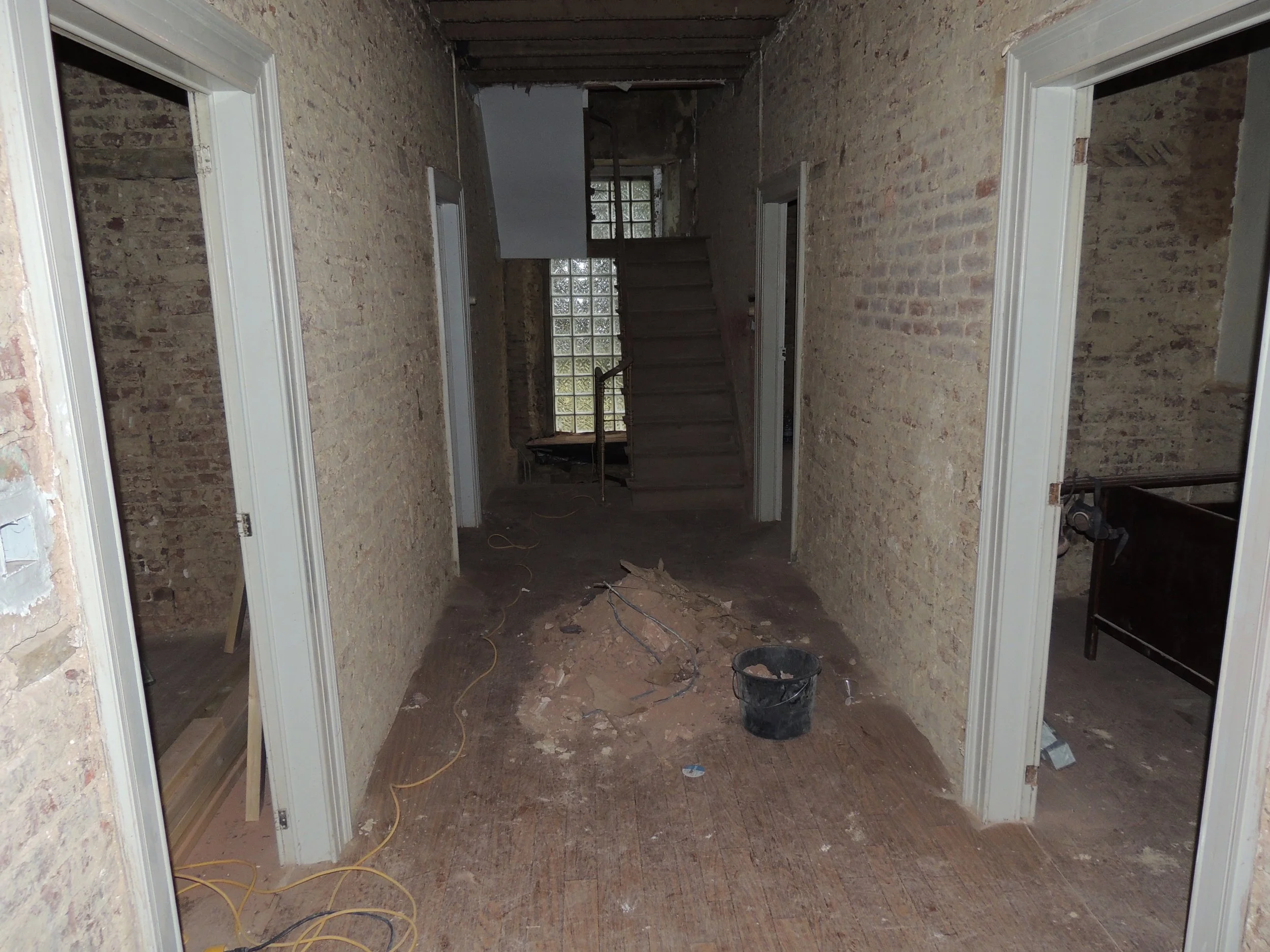 Intérieur en cours de rénovation avec murs en briques, absence de plafond, sol en parquet partiel, et fenêtres en verre de bloc.