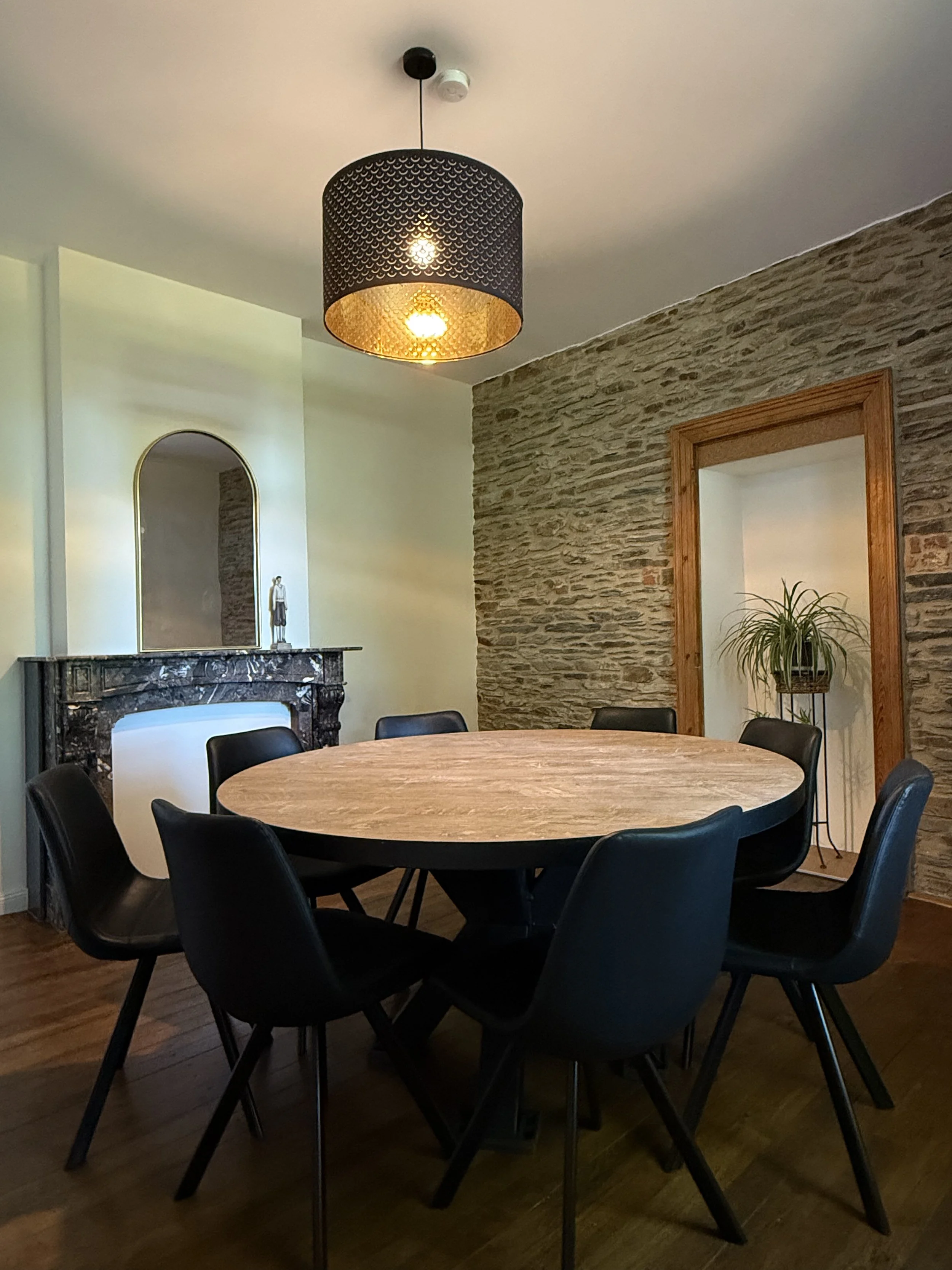 Salle à manger avec une table ronde en bois, six chaises en plastique noir, un mur en pierre décorative, un miroir encadré en bois, une plante en pot et un lampadaire suspendu noir et doré.