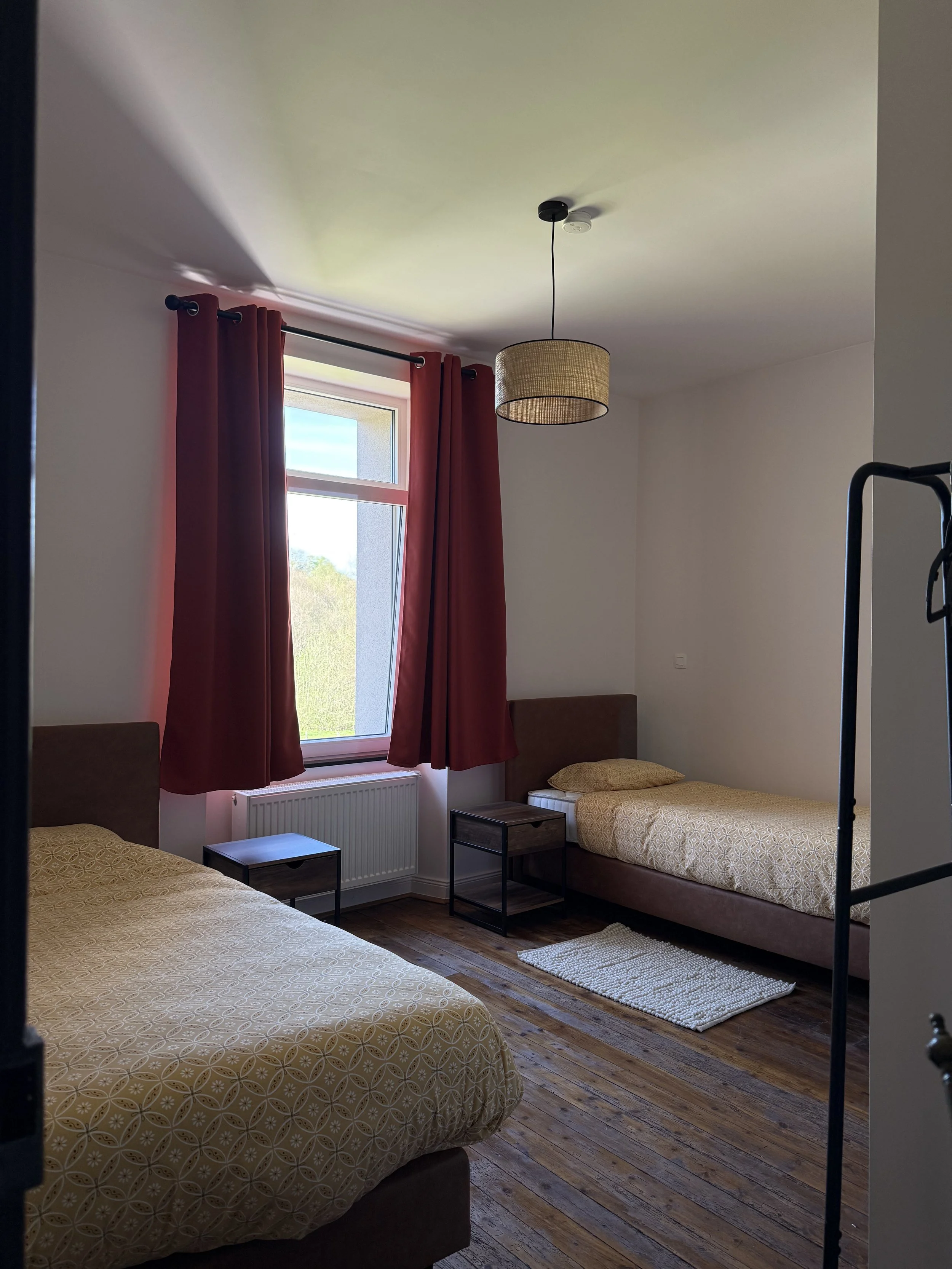 Chambre avec deux lits jumeaux à côté d'une fenêtre avec rideaux rouges, deux tables de chevet en bois, un tapis blanc, un plafond avec un luminaire en suspension, et des murs blancs avec un sol en bois.