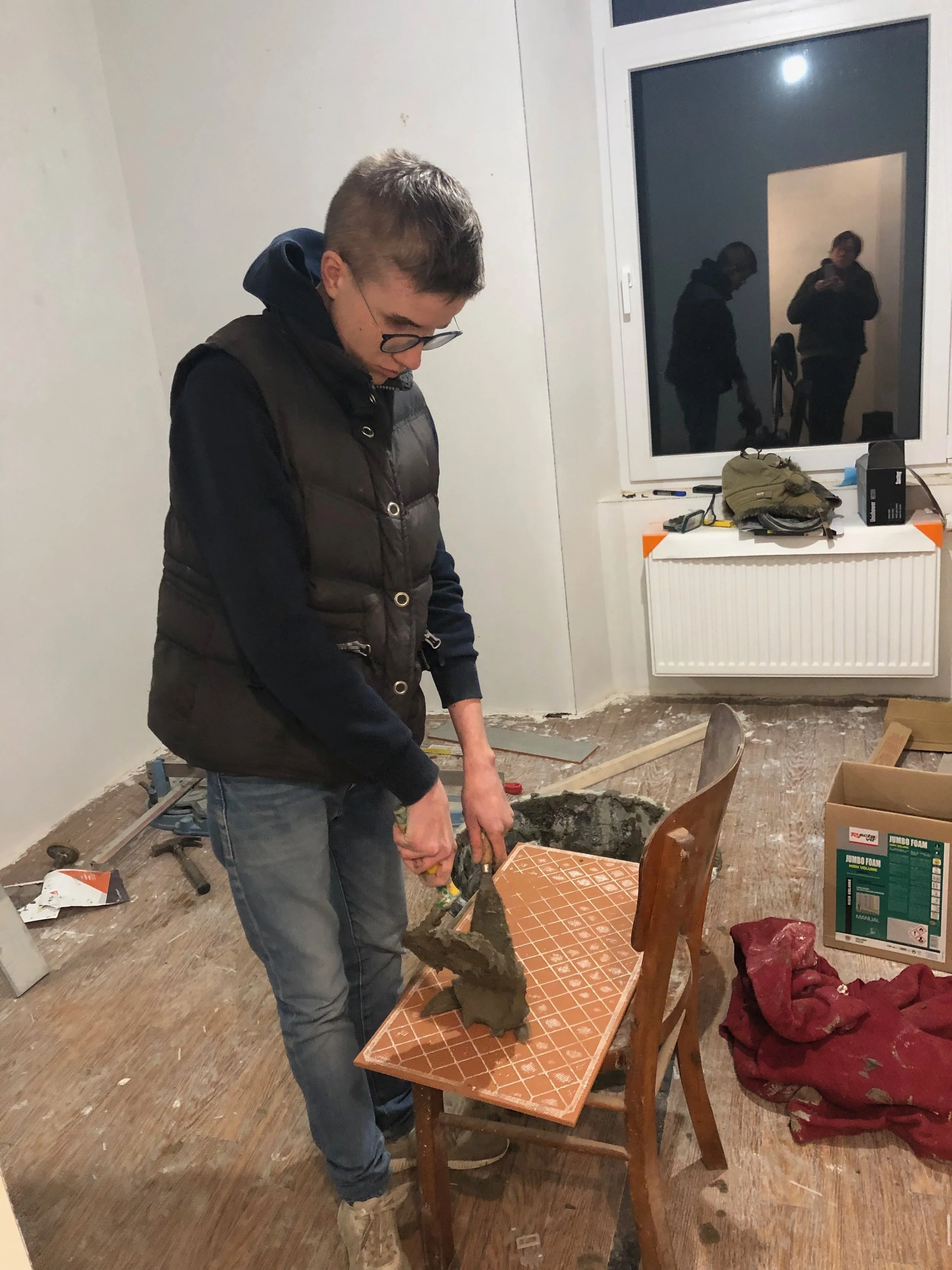 Un jeune homme en vêtements décontractés travaille dans une pièce en rénovation, debout à côté d'une table sur laquelle il manipule un objet. Il porte des lunettes et un gilet. La pièce a un sol en bois, une fenêtre, et des outils et matériaux de construction partout, notamment un sac de bricolage, des cartons, et du matériel poussiéreux.