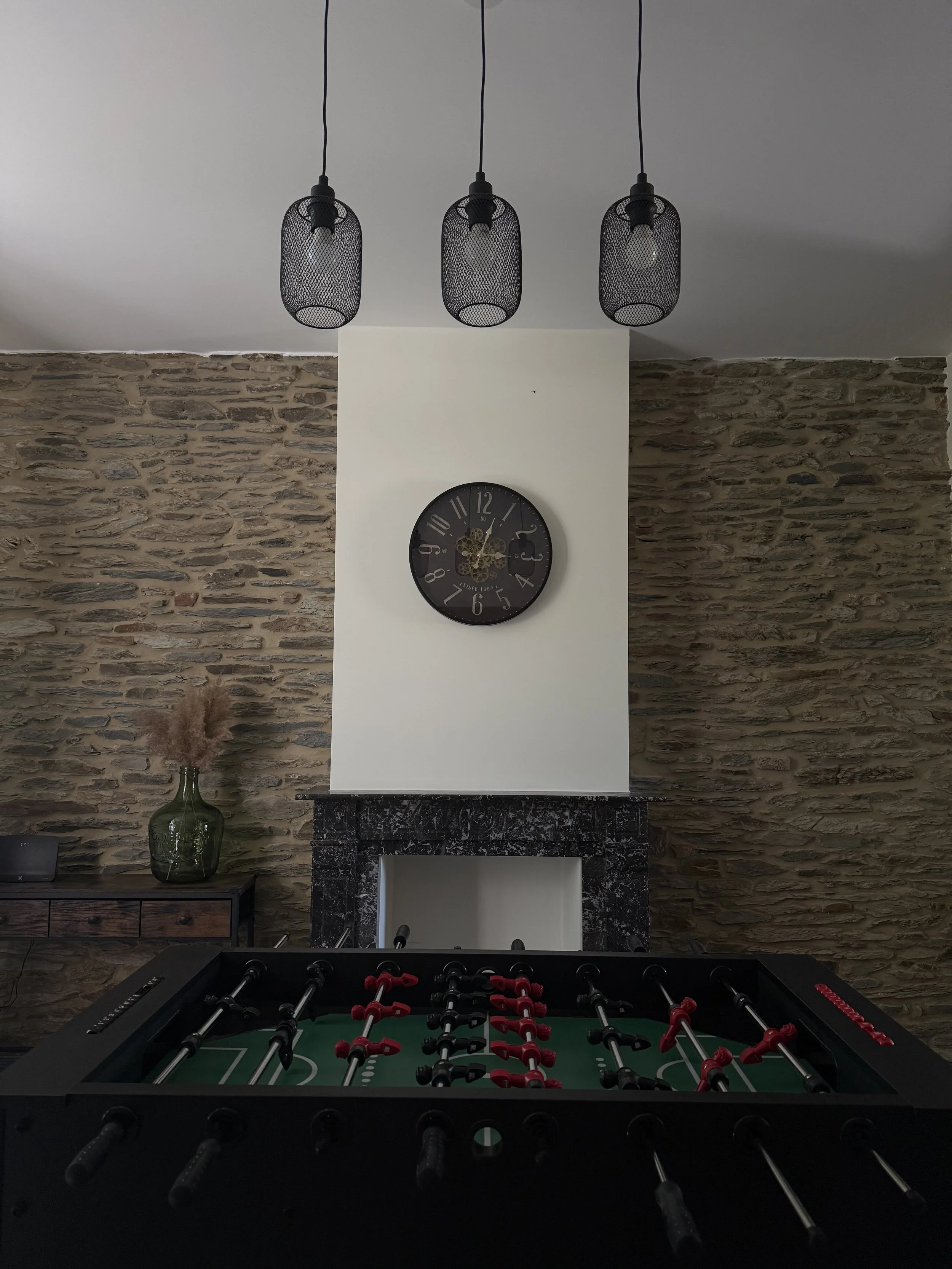Au premier plan, un babyfoot avec des joueurs rouges et noirs. En arrière-plan, un mur en pierre et un meuble en bois avec une grande bouteille en verre contenant des végétaux. Au centre, un mur avec une horloge murale noire. Au plafond, trois lampes noires suspendues.