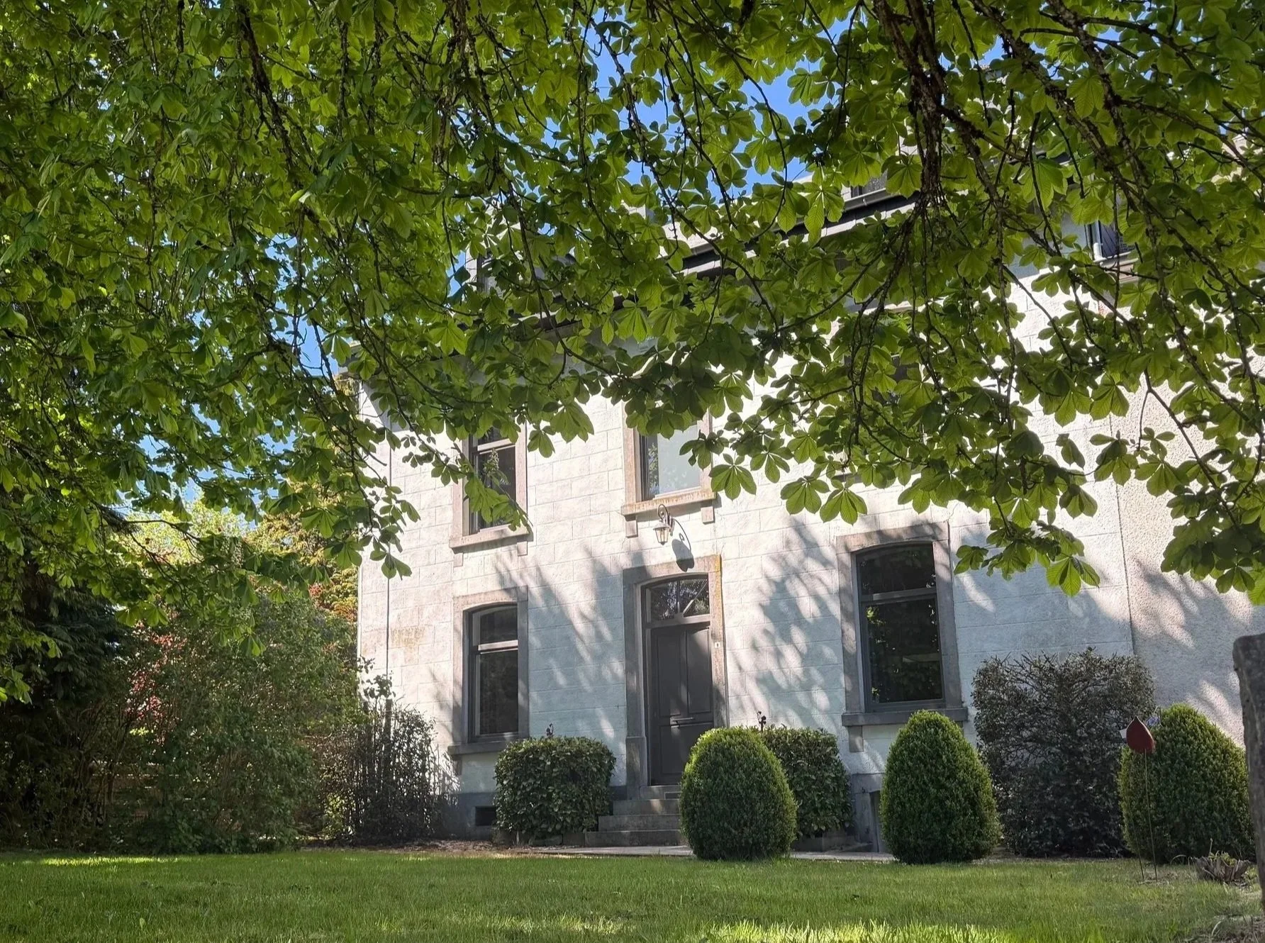 Maison en pierre avec un jardin bien entretenu, de grands arbres à feuilles vertes, et un ciel bleu.