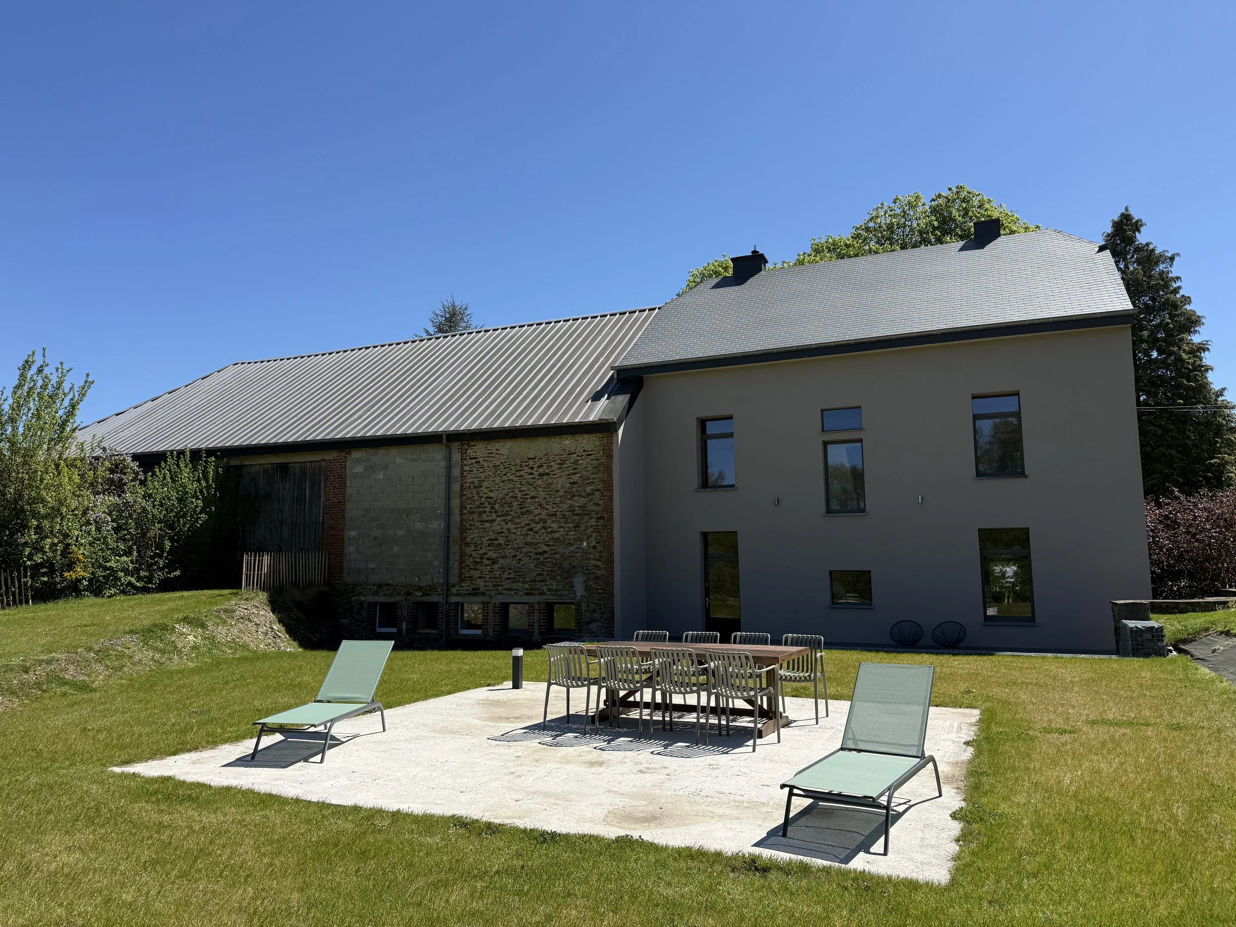 Une maison moderne avec une toiture en tuiles grises, entourée d'un jardin vert. À l'avant, il y a une terrasse avec des chaises longues en plastique vert et une table avec des chaises. Le ciel est clair et bleu, sans nuages.
