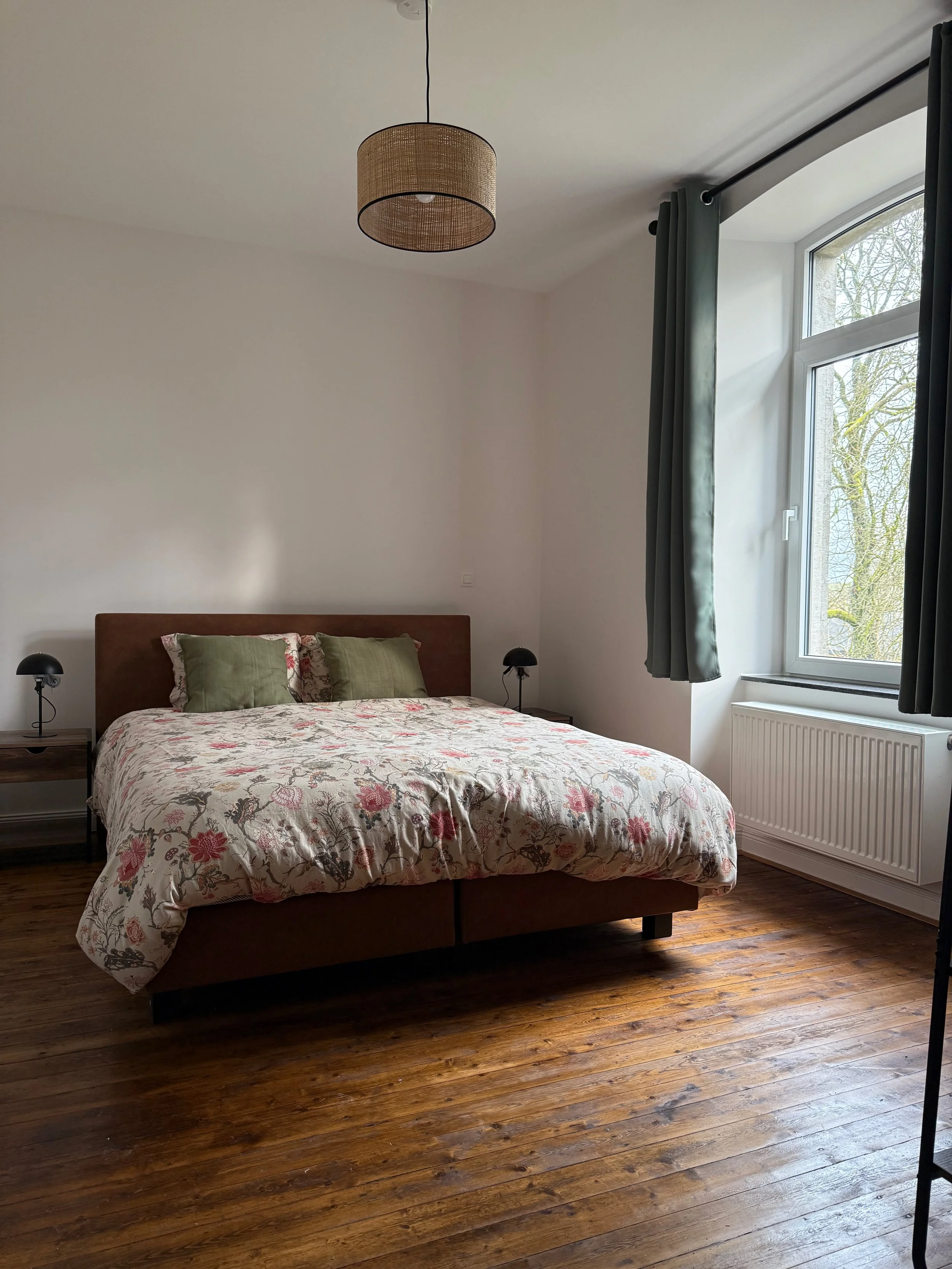 Chambre avec un lit à tête en bois, drap à motifs floraux, coussins verts, lampes de chevet de chaque côté, fenêtre avec rideaux gris foncé, plancher en bois.