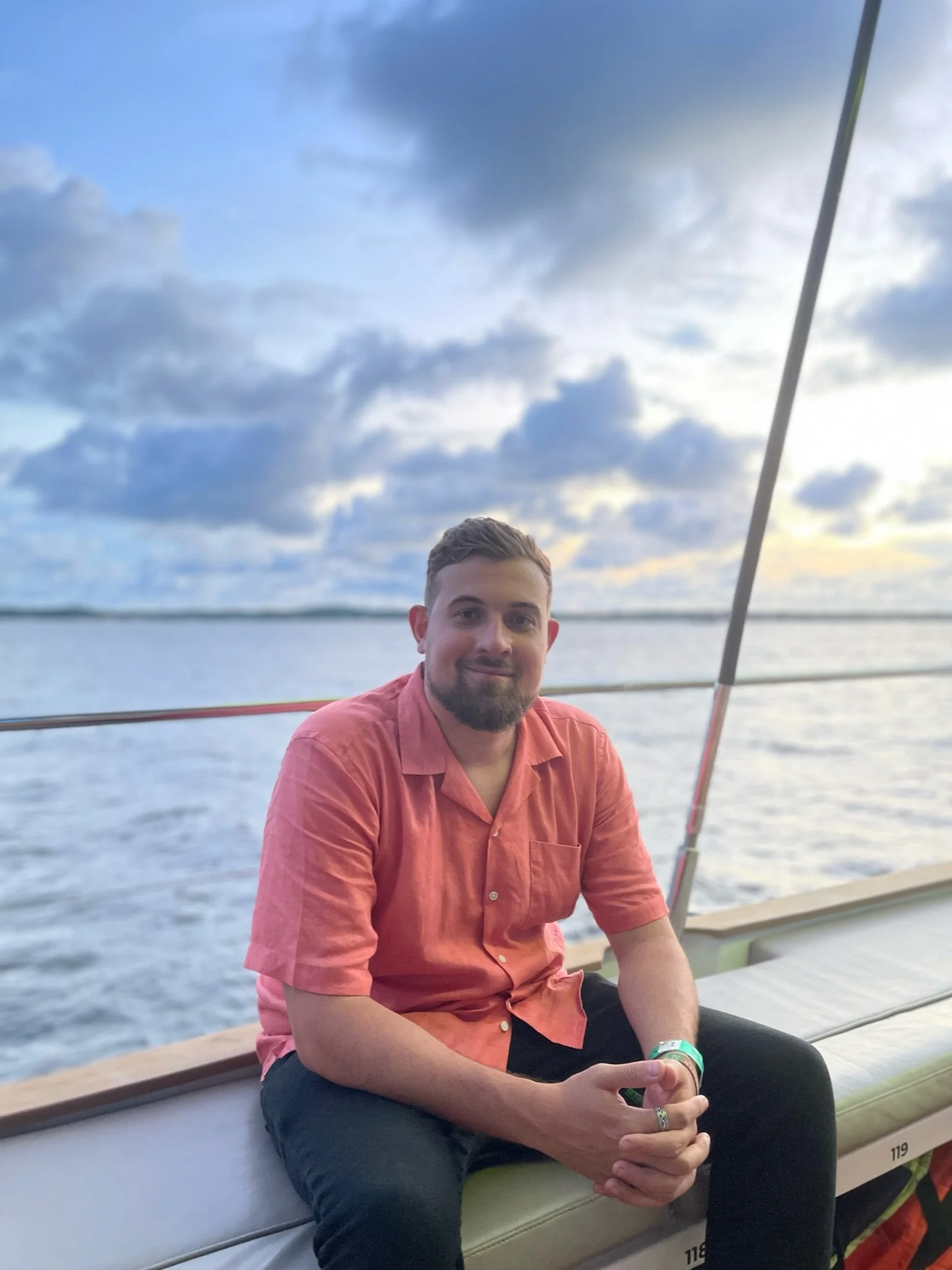 A man sitting outdoors on a boat with a lake and cloudy sky in the background, smiling at the camera wearing a salmon-colored shirt and black pants.