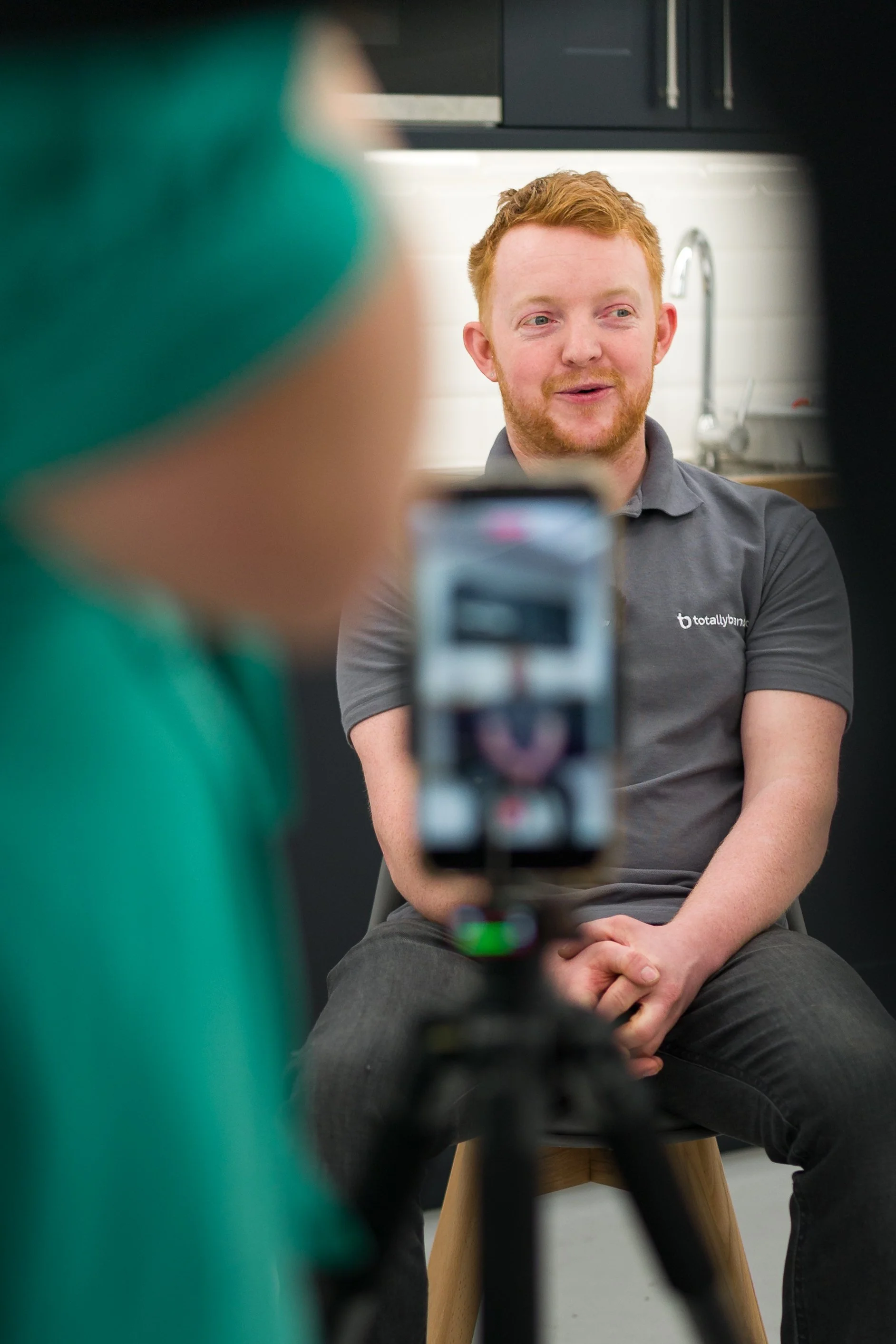 A red-haired man with a beard, wearing a gray shirt, is sitting on a stool with his hands folded on his lap. He is smiling slightly and looking to his right, while a person in green is in the foreground taking a photo with a smartphone. The background shows a kitchen with black cabinets, a white tiled backsplash, and a stainless steel sink.
