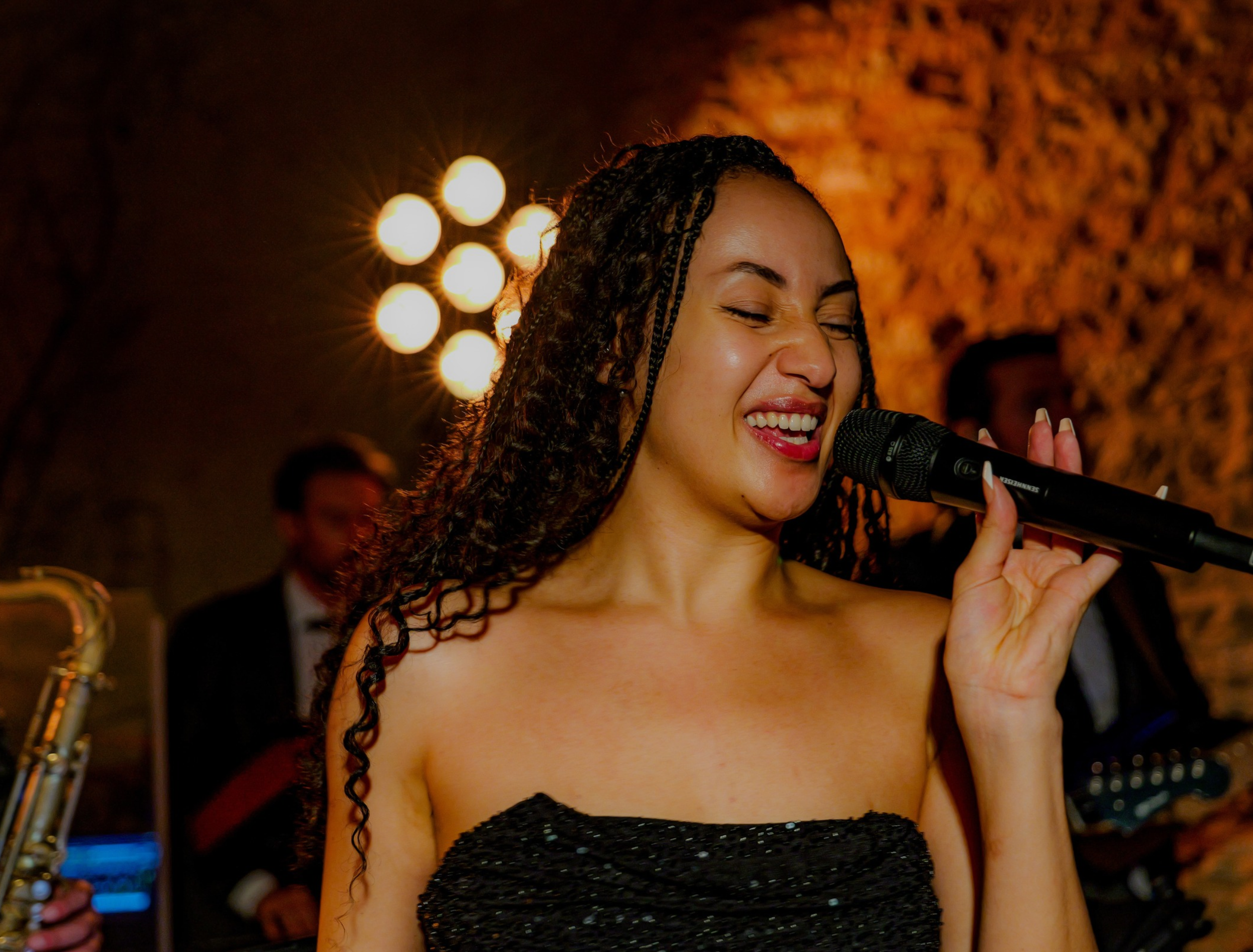 A woman singing into a microphone with closed eyes and a smile, at a live music performance, with stage lights in the background.