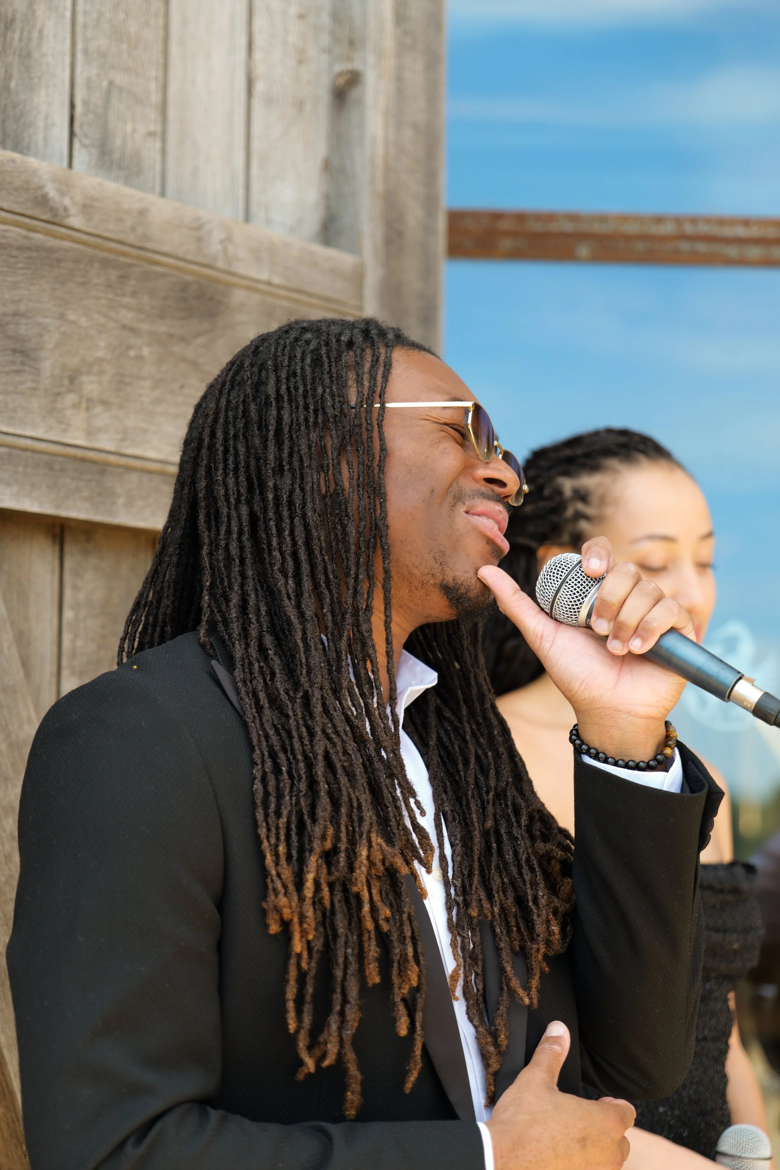 A man with long dreadlocks, wearing sunglasses and a black suit, is holding a microphone and singing or speaking. Behind him, part of a woman's face and hand are visible, and she appears to be singing or speaking. They are outdoors with a wooden structure and blue sky in the background.