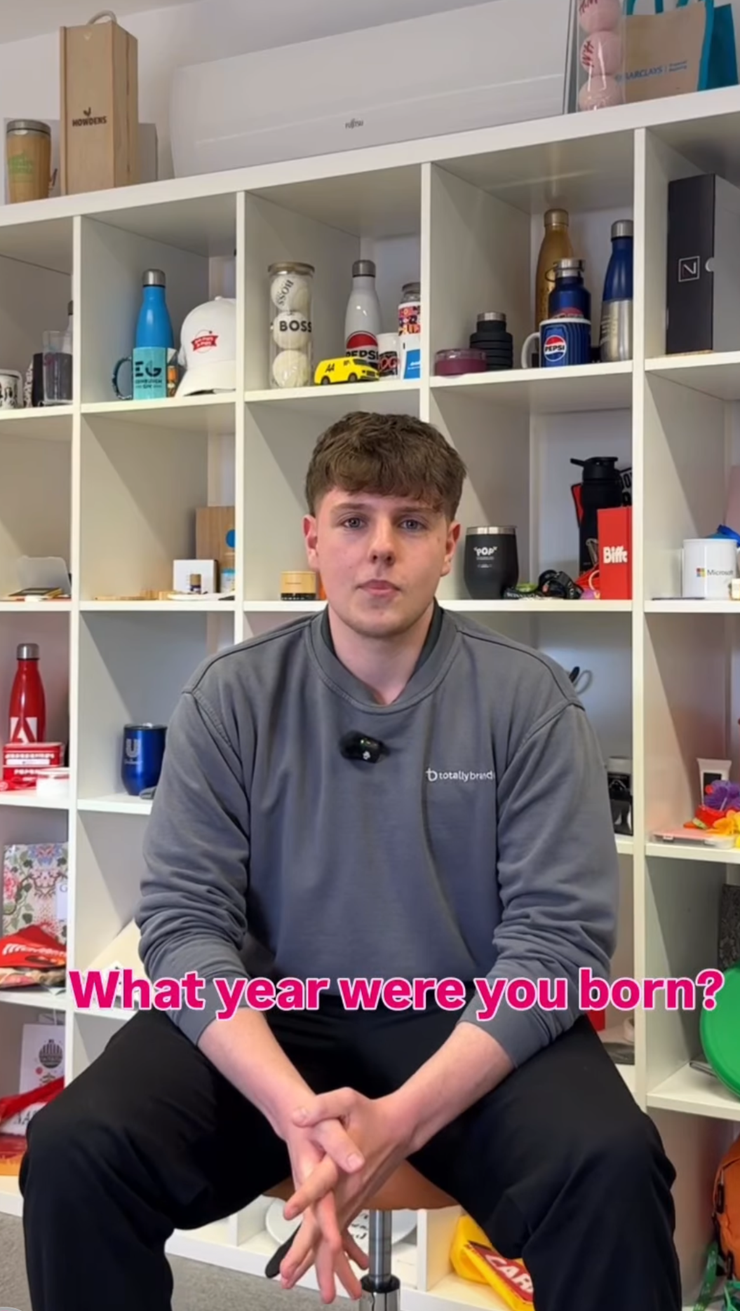 A young man with brown hair sitting in front of a white cubical shelf filled with various objects including bottles, jars, and accessories. He is wearing a gray sweatshirt and black pants, and is looking directly at the camera with a neutral expression.