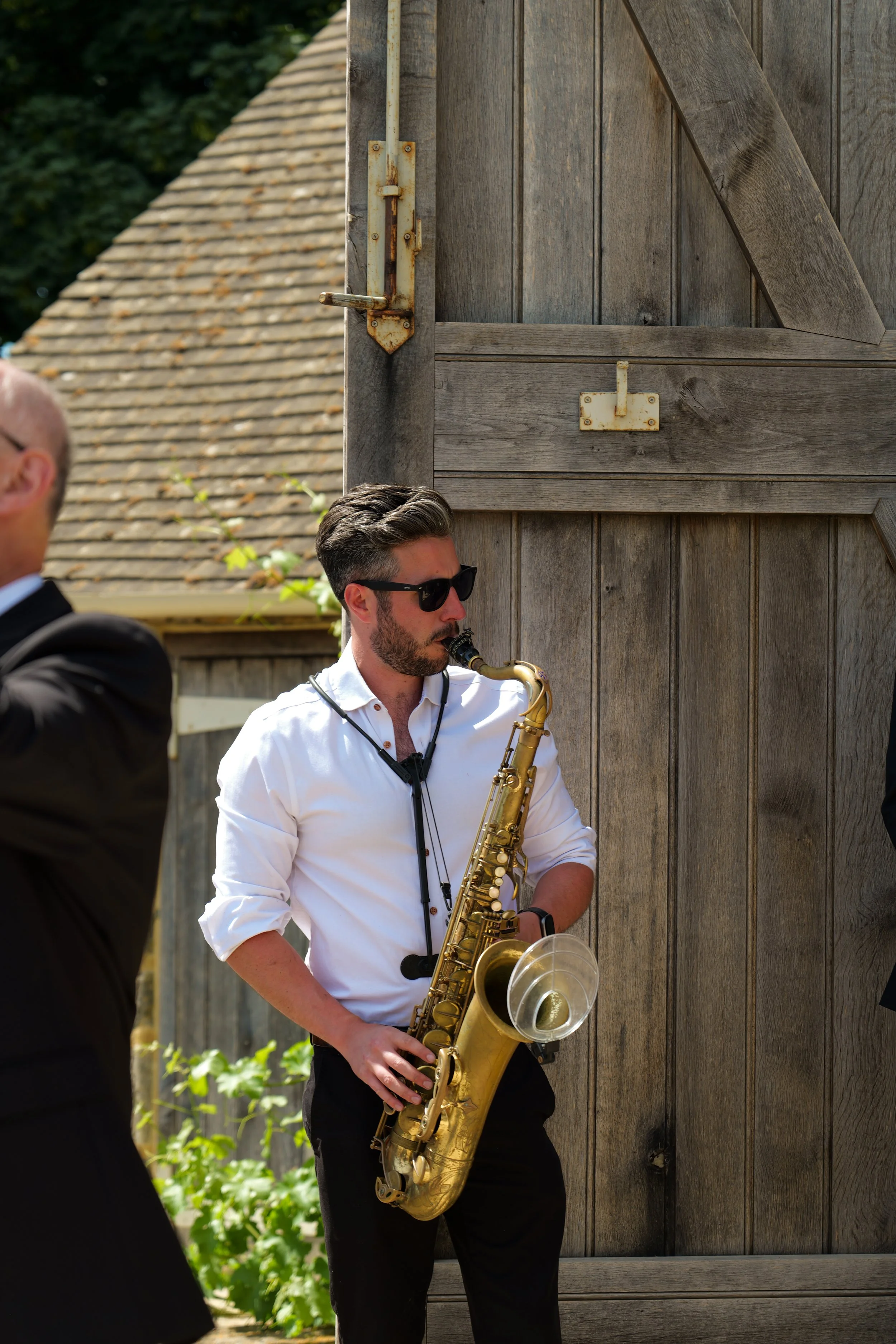 A man in white shirt wearing sunglasses playing a saxophone outdoors, standing next to a wooden door and a person in black suit in the foreground, with a house and greenery in the background.