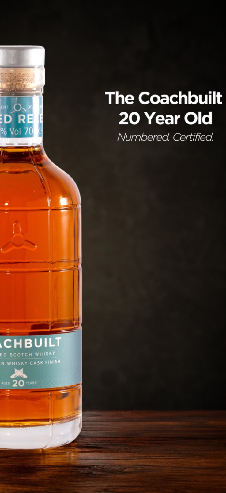 Close-up of a bottle of The Coachbuilt 20 Year Old Scotch Whisky on a wooden surface, with a dark background and white text to the right describing the product as aged, numbered, and certified.