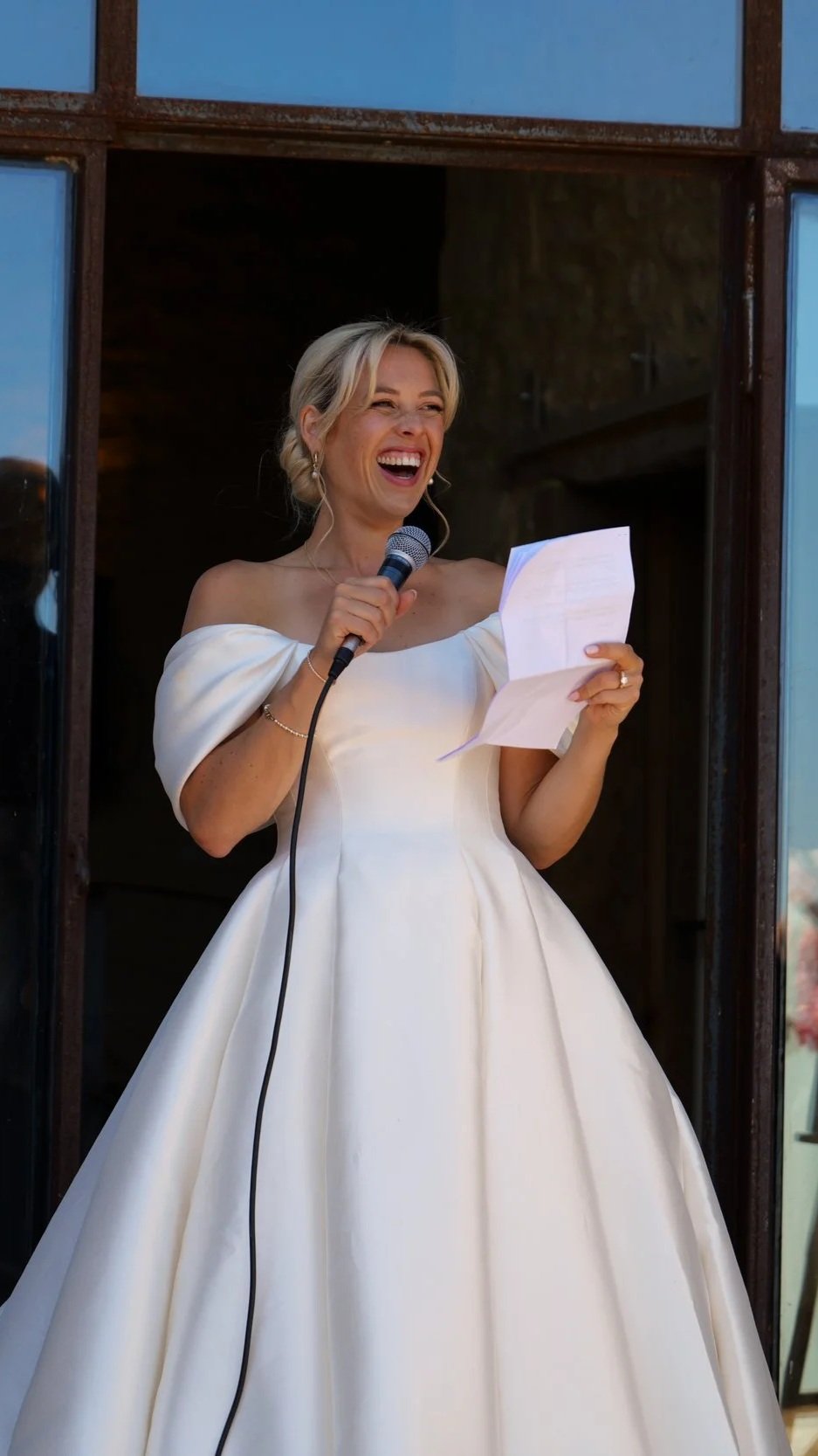 A woman in a white off-shoulder gown is smiling and speaking into a microphone while holding a piece of paper, standing in front of a rustic building window.