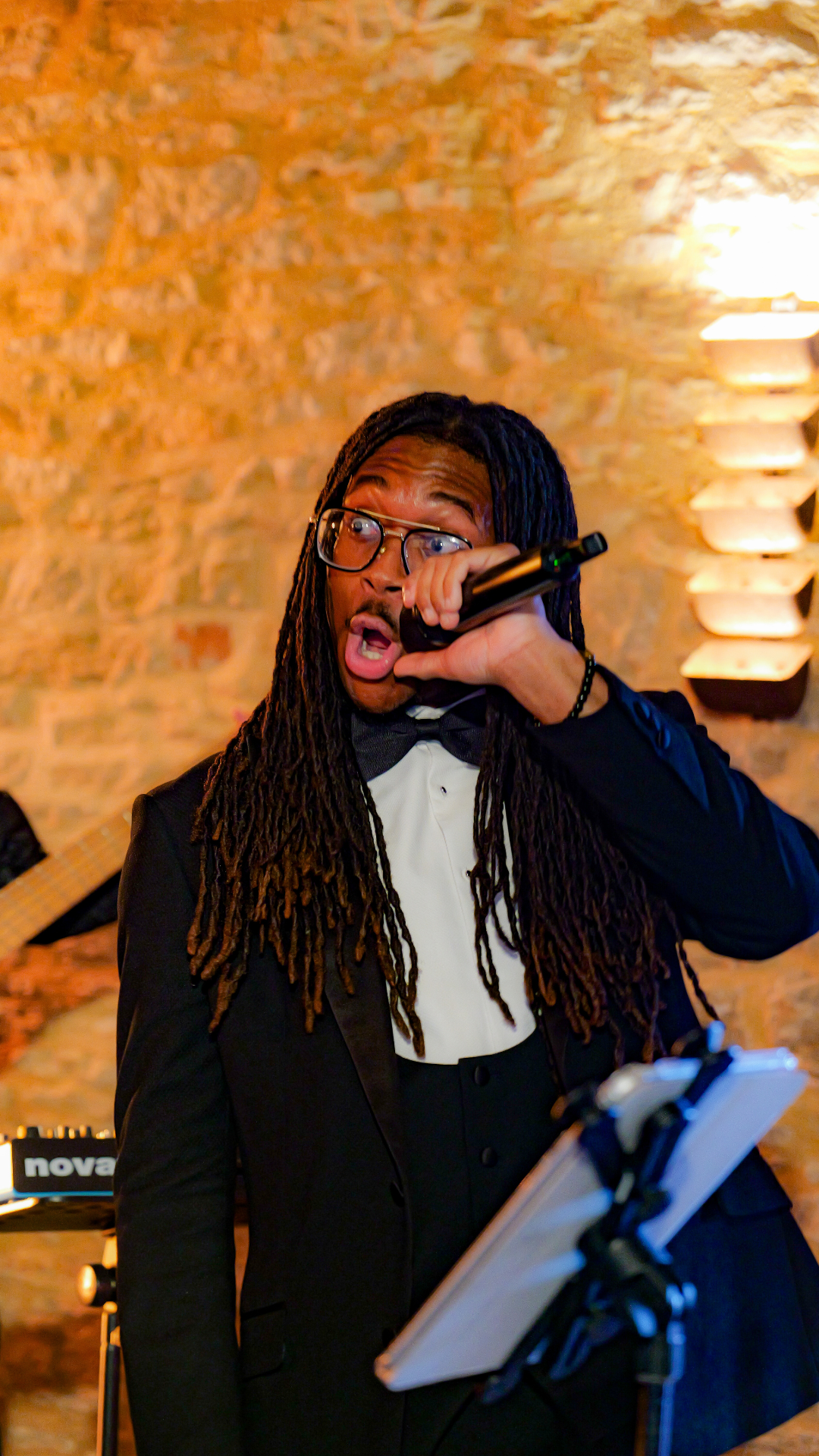 A man with long dreadlocks, glasses, and a tuxedo singing into a microphone at a performance venue with brick walls and warm lighting.