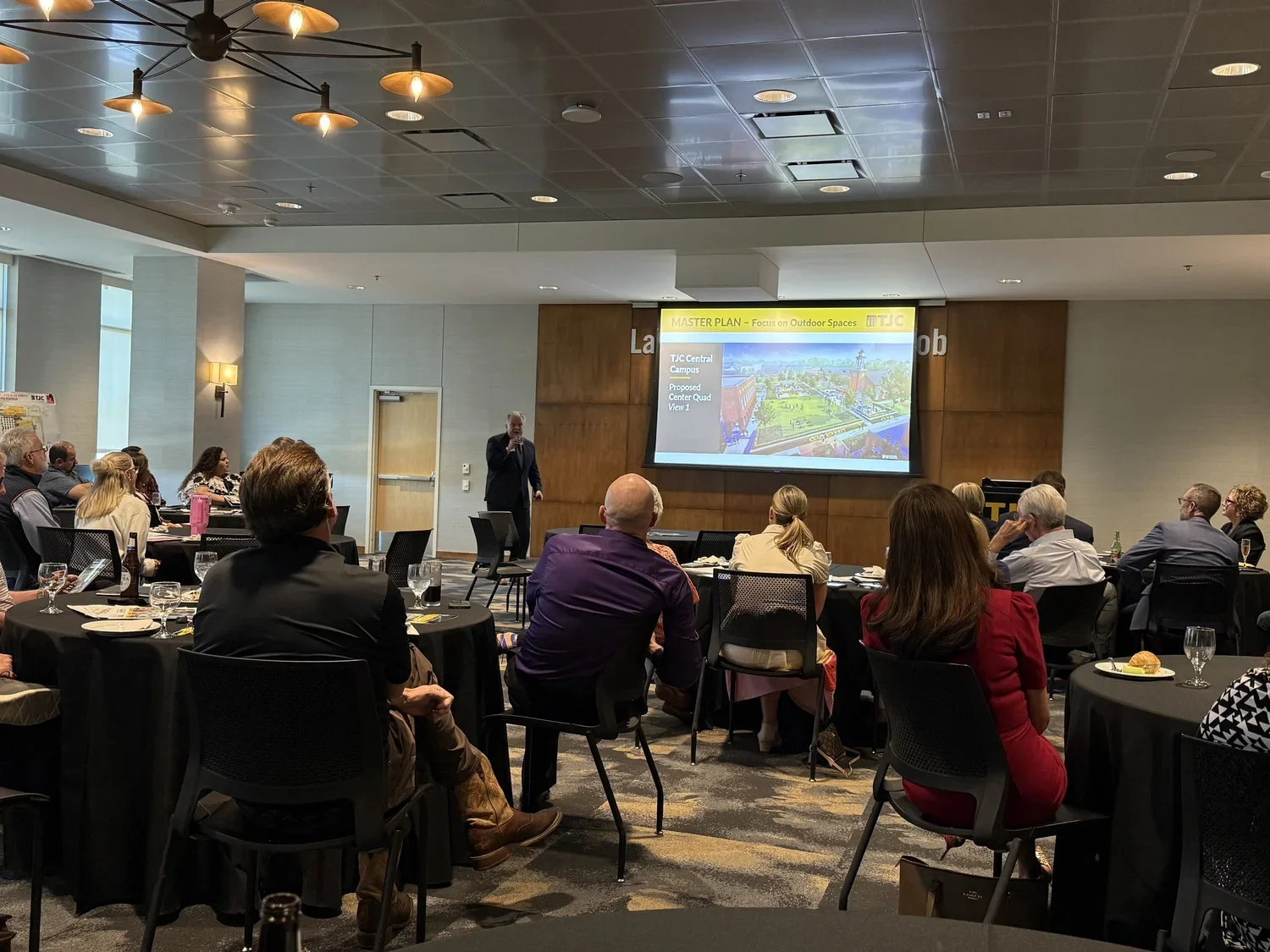 Photo of a meeting room with attendees seated at round tables while a presenter stands at the front near a projected screen.