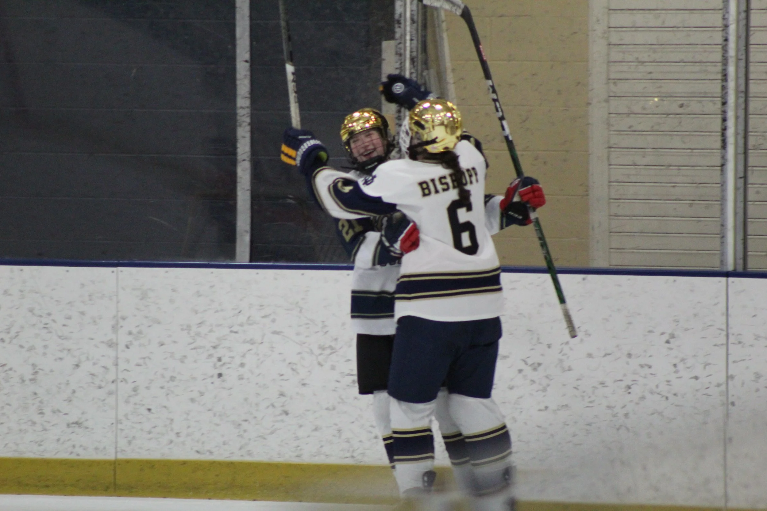 Two girls in hockey uniforms and gold helmets celebrating on the ice rink, with one girl holding a hockey stick.