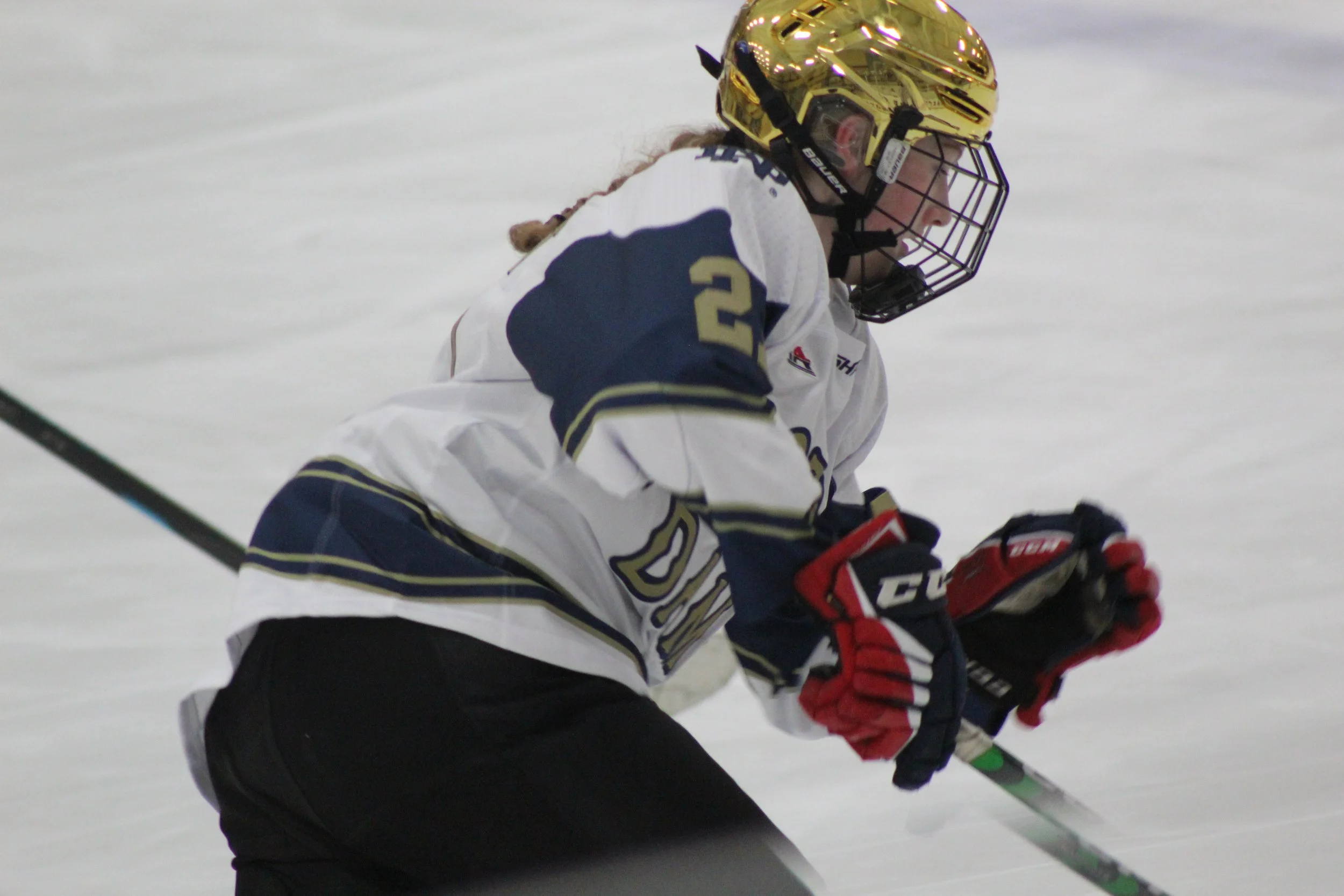 Hockey player in a white jersey with navy blue and gold accents, wearing a gold helmet with a face cage, skating on ice with a hockey stick, during a game.
