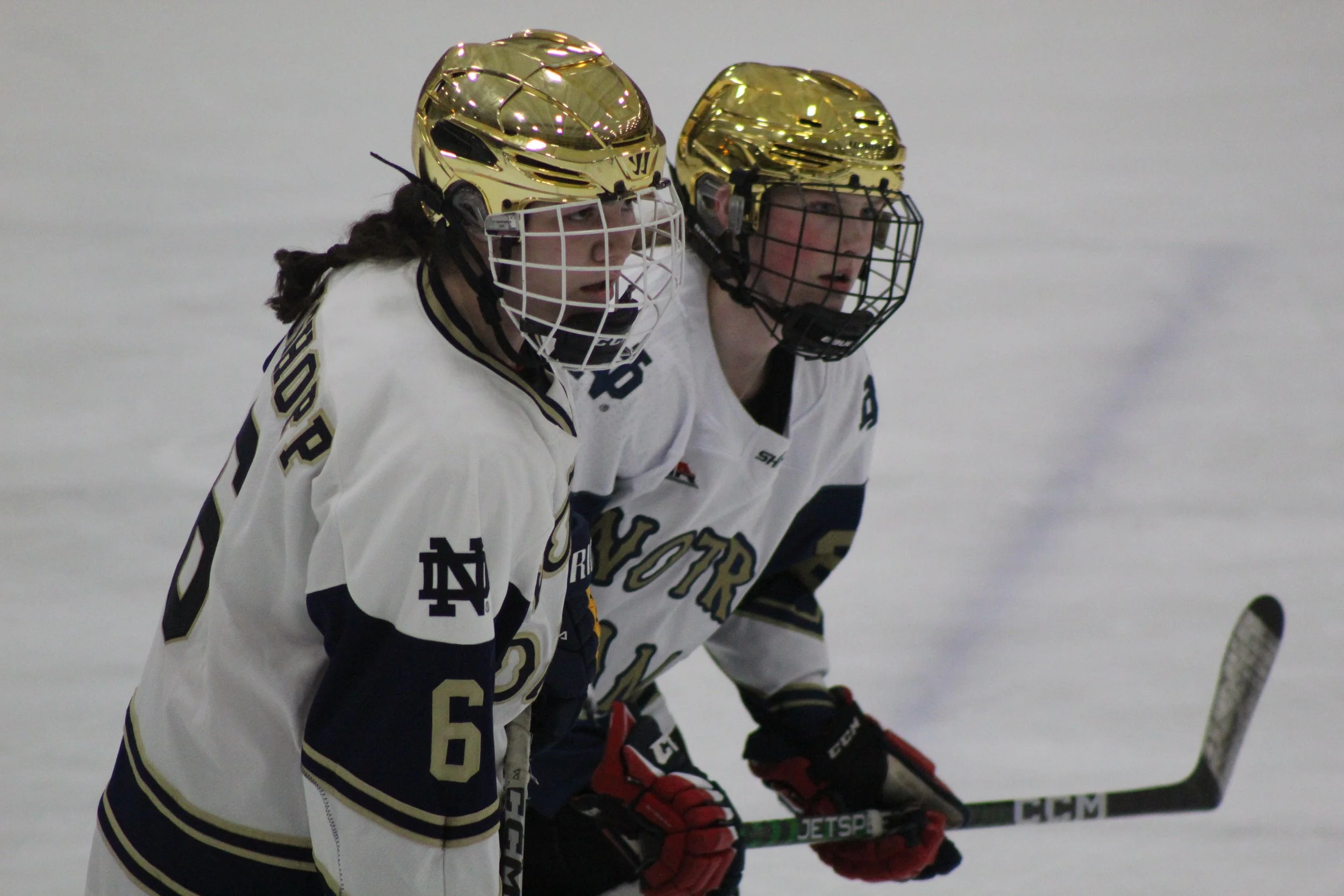 Two female ice hockey players wearing white jerseys with navy and gold details, gold helmets, and protective gear, are standing close together on the ice during a game.