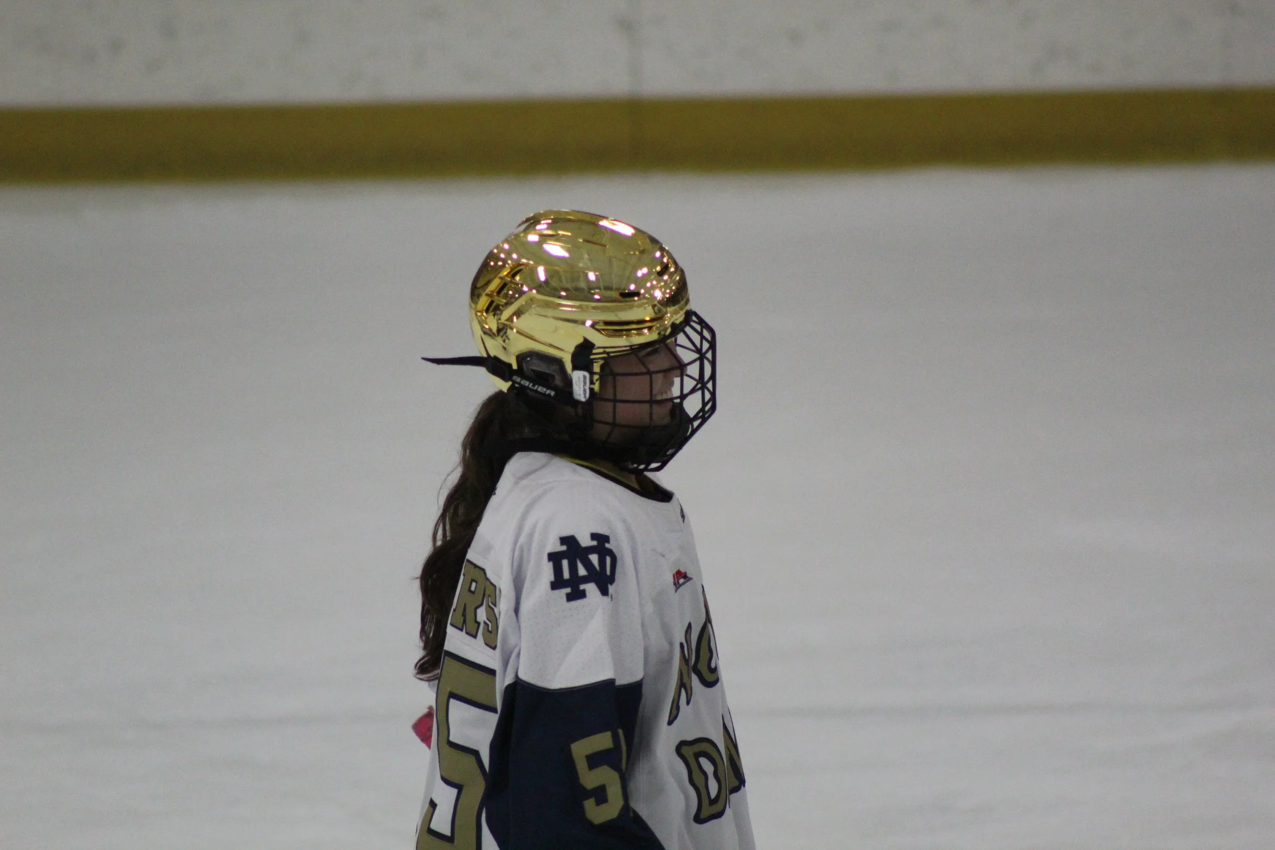 An ice hockey player on the ice wearing a white and navy jersey with the number 5, a black helmet with a gold visor, and a face cage, appears to be looking forward.