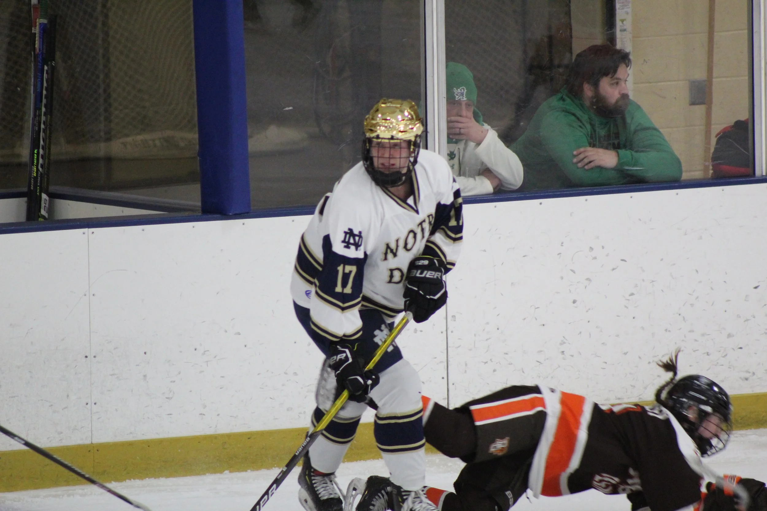 Hockey player in a white jersey with number 17 skating on ice during a game, with a fallen player in a black and orange jersey nearby, and spectators watching through the glass in the background.