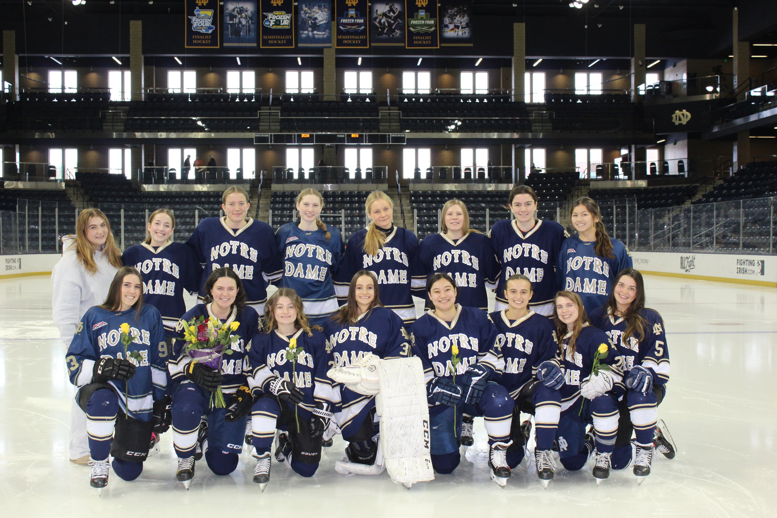 A women's ice hockey team in matching blue jerseys with 'Notre Dame' written on them, posing on an ice rink inside an indoor arena with banners hanging overhead.