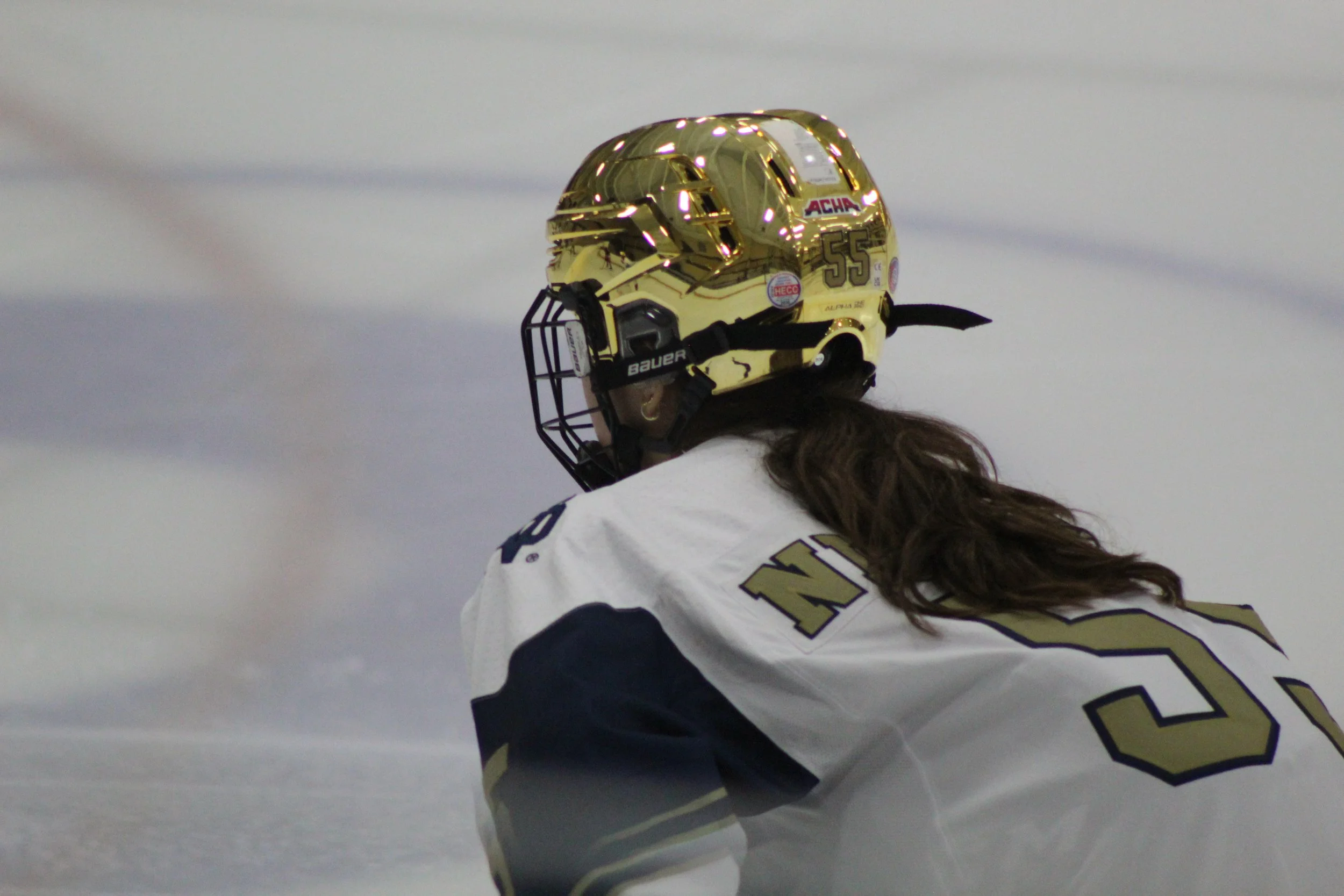 Ice hockey player wearing a gold helmet and white jersey with navy accents, kneeling on the ice during a game.