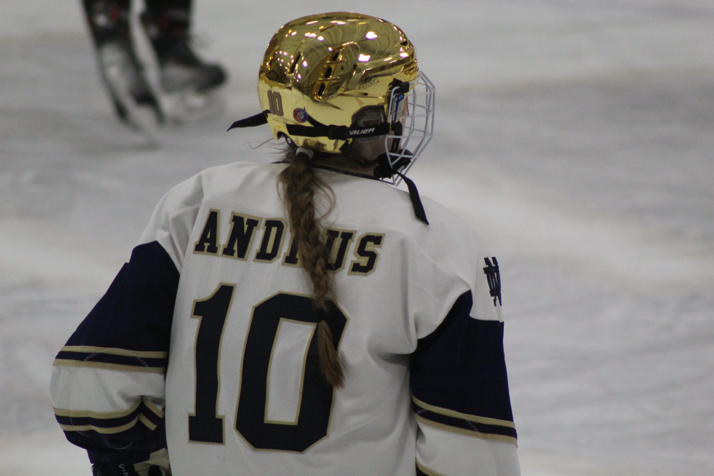 Ice hockey player wearing a white jersey with the name 'ANDUS' and the number 10, on an ice rink, with a shiny gold helmet, and a braid hanging down the back.
